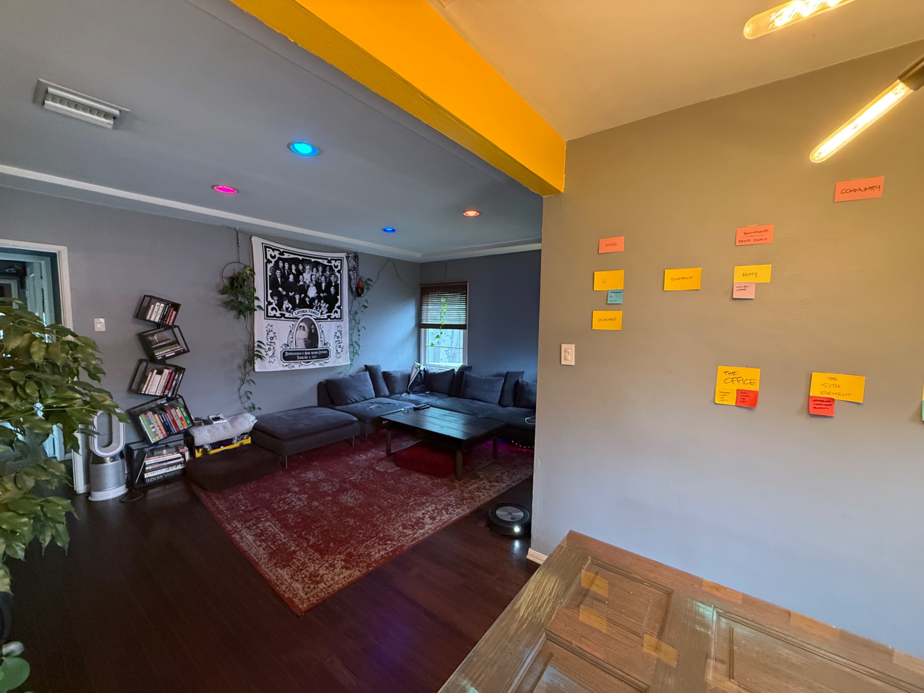 The room features a contemporary layout with a gray color scheme, a large wall tapestry, a seating area with two sofas, and brightly colored sticky notes affixed to a nearby wall.