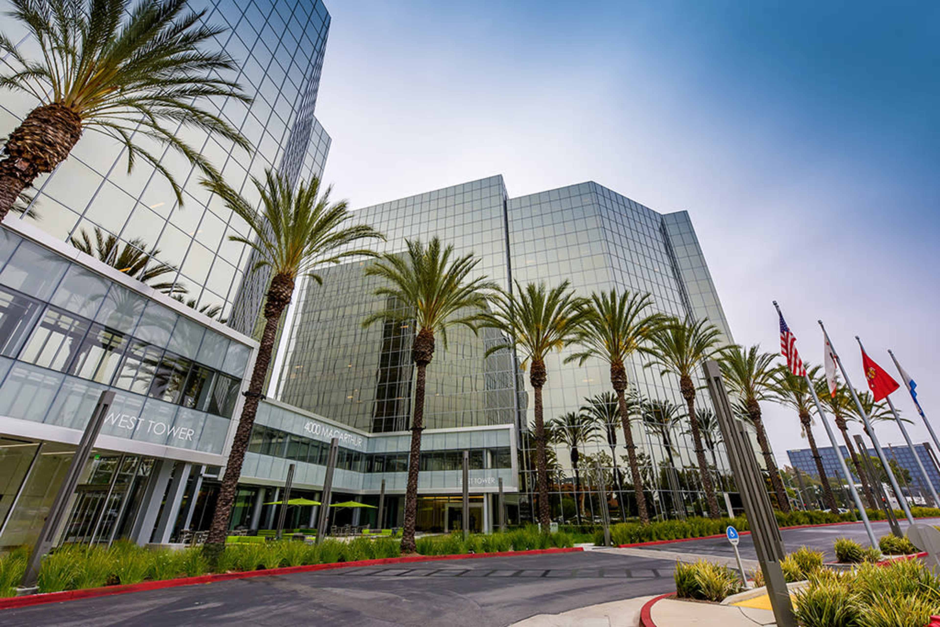 The image shows a modern office building with large glass windows, palm trees lining the driveway, and several flags displayed in front.