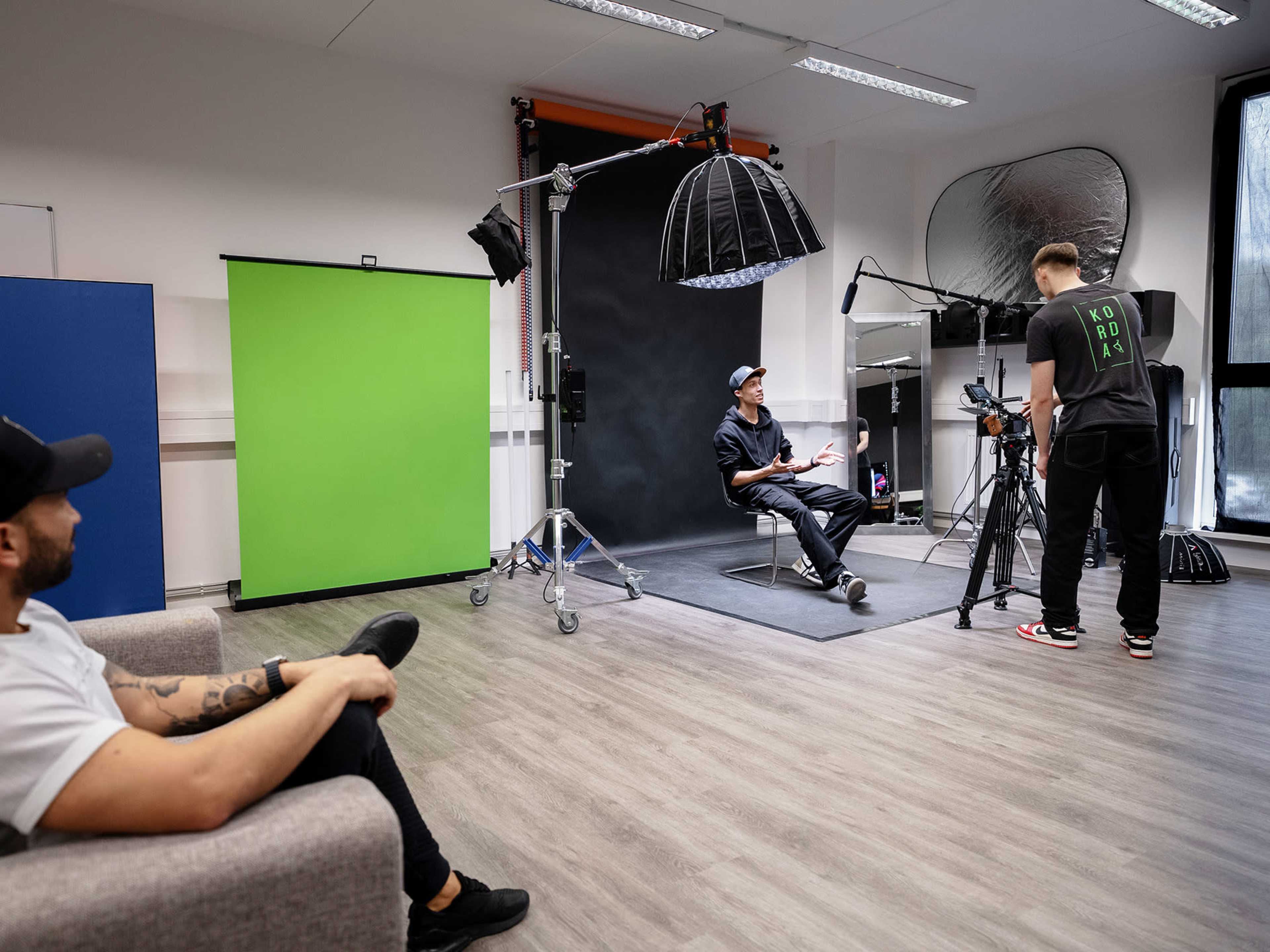 A person in a chair is being filmed in a studio set up with a green screen and various lighting equipment.