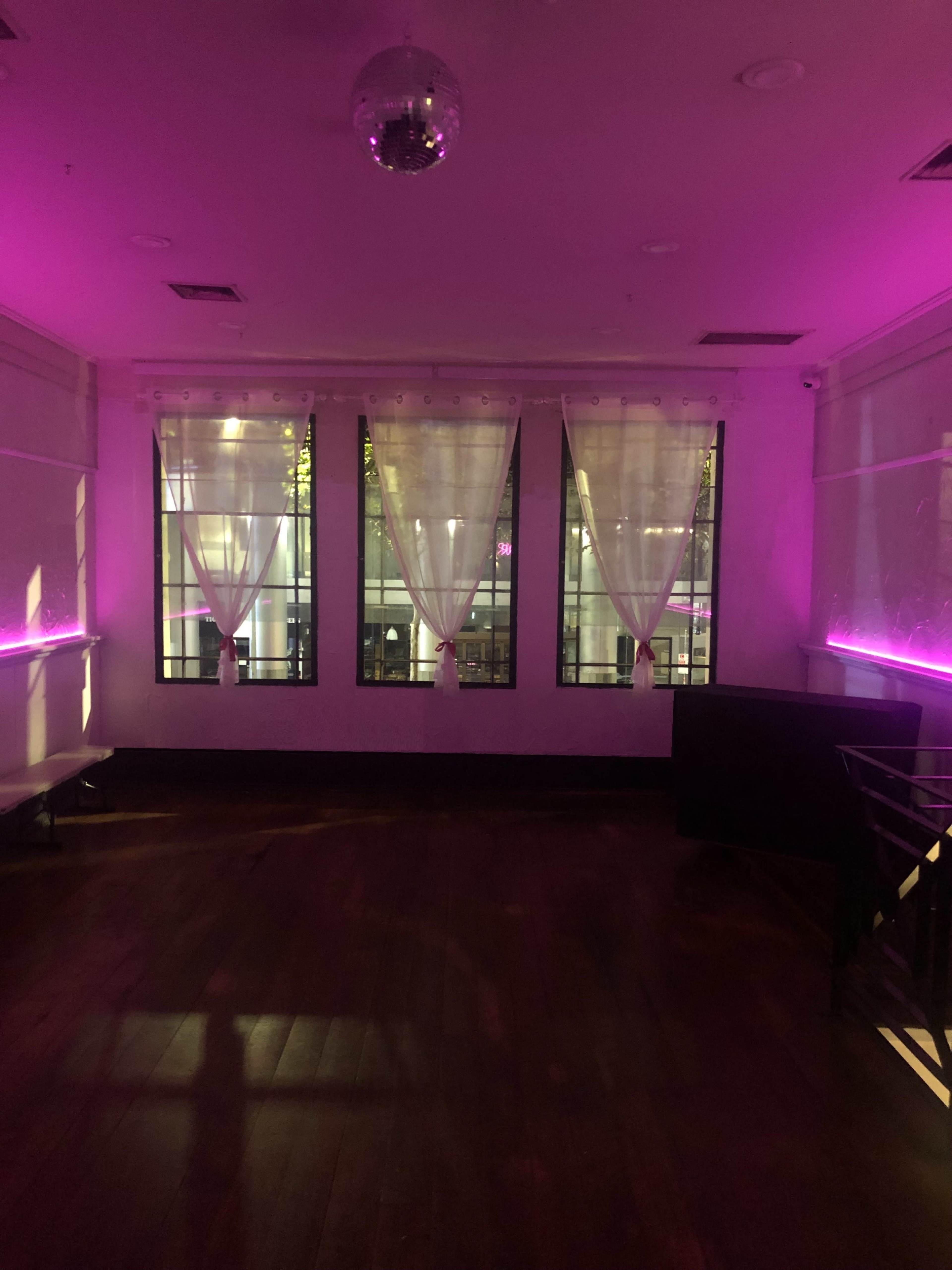 The image shows a dimly lit room with purple lighting, large windows, sheer white curtains, and wooden flooring.