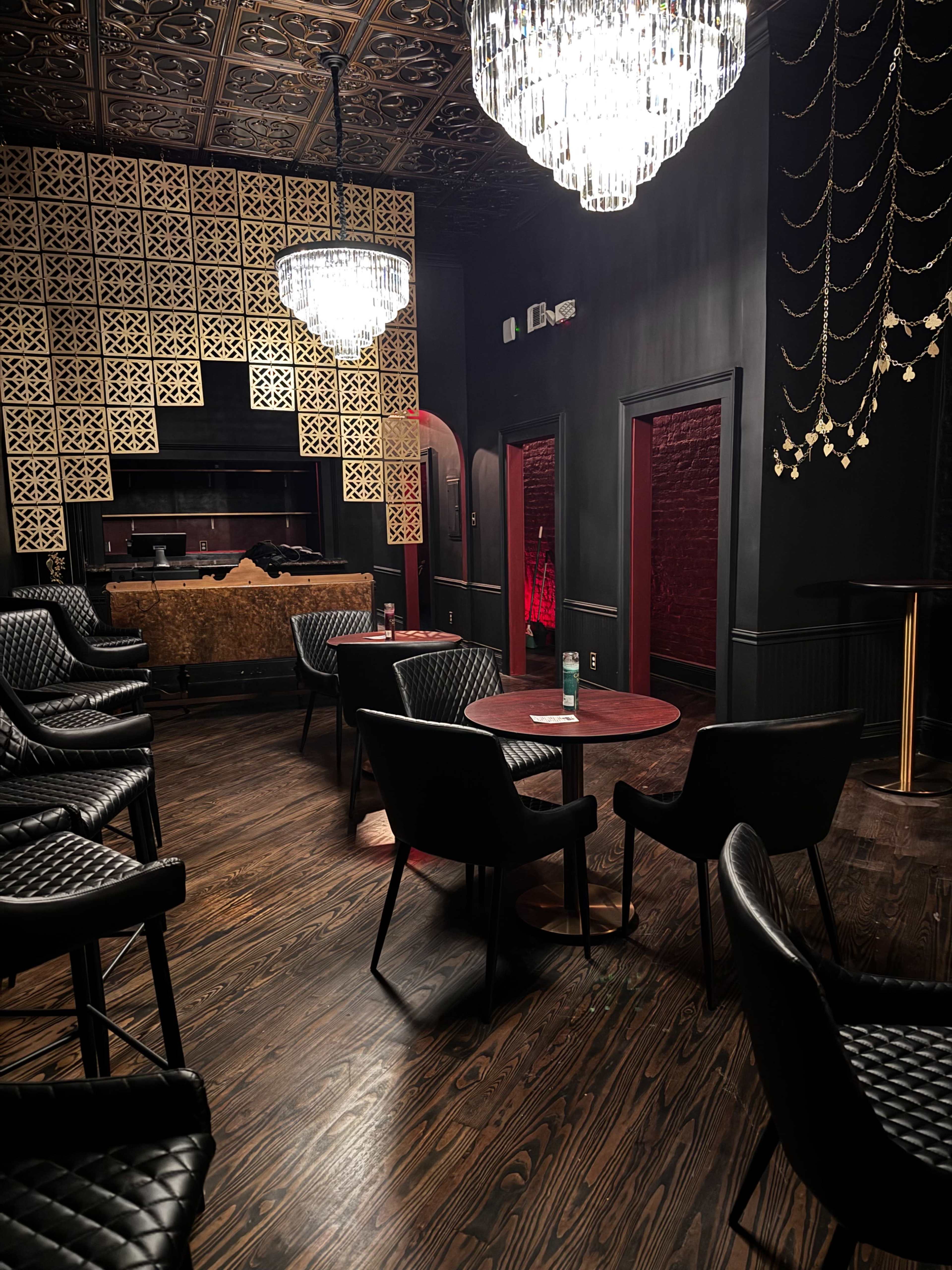 The image shows a dimly lit lounge with dark walls, ornate ceiling tiles, crystal chandeliers, and black chairs arranged around a small table.