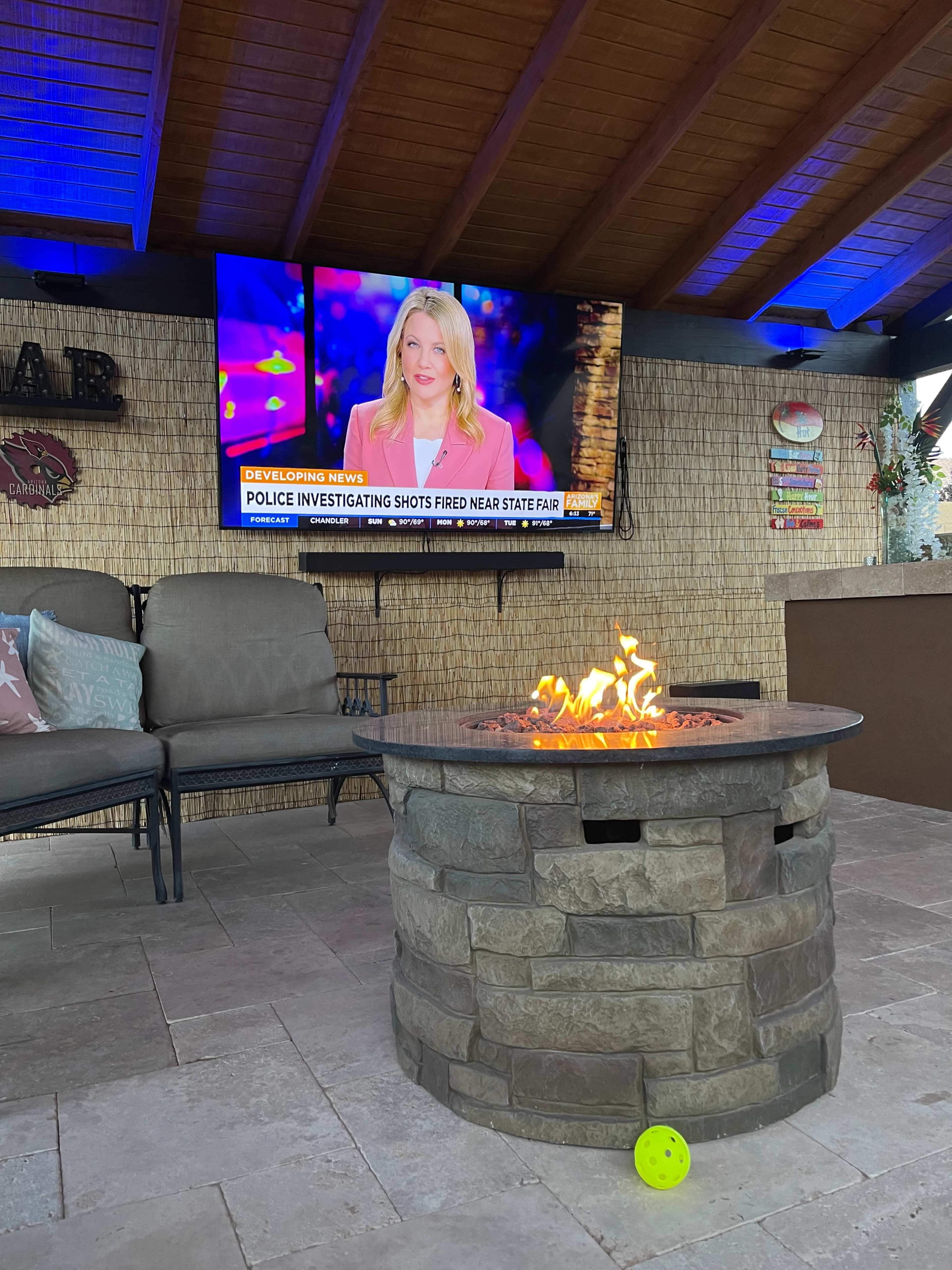 A stone fire pit with a flame is in the foreground, while a television broadcasts news about police investigating shots fired near a state fair in the background.