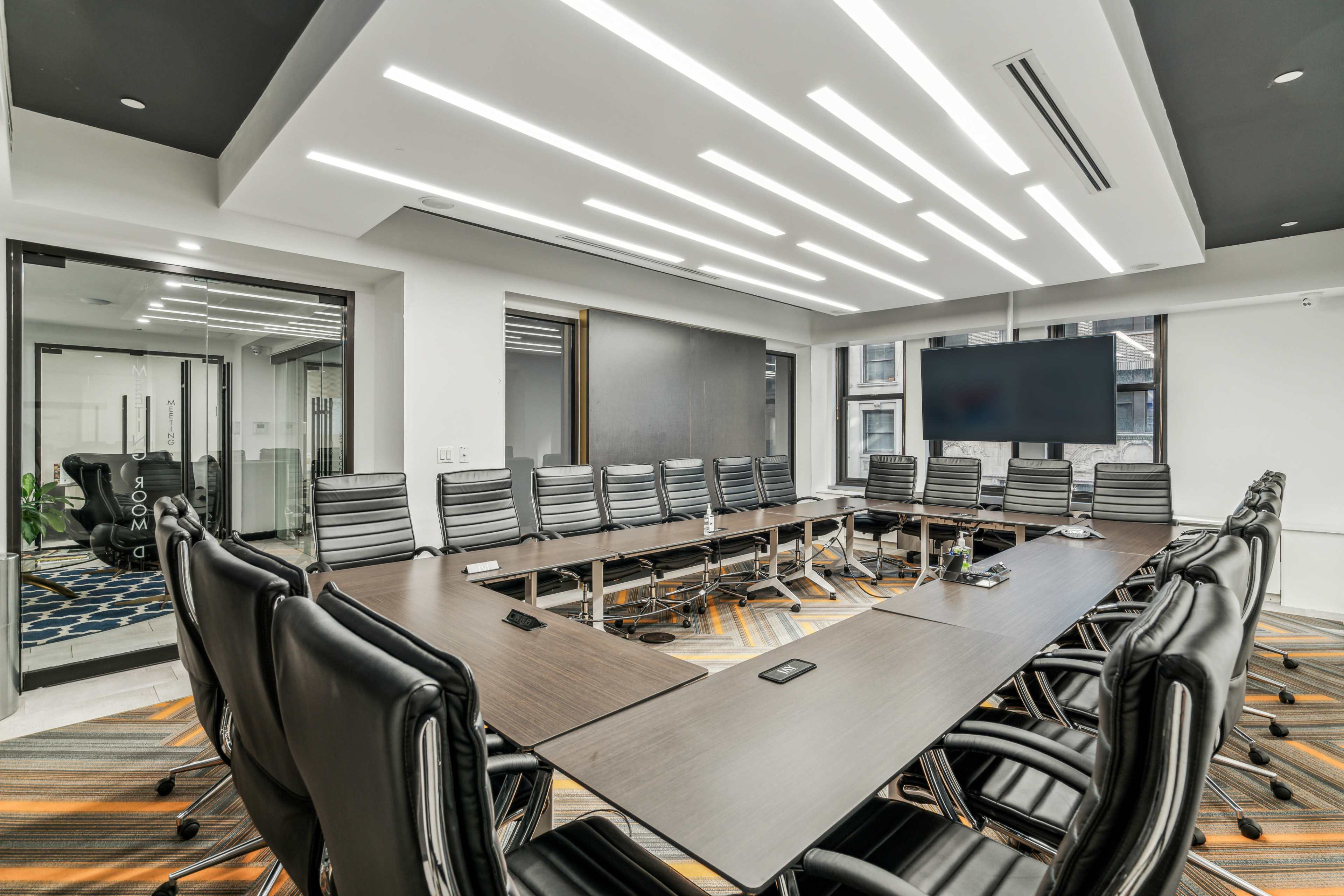 A modern conference room features a large rectangular table surrounded by black leather chairs, illuminated by unique ceiling lights and equipped with a large wall-mounted screen.