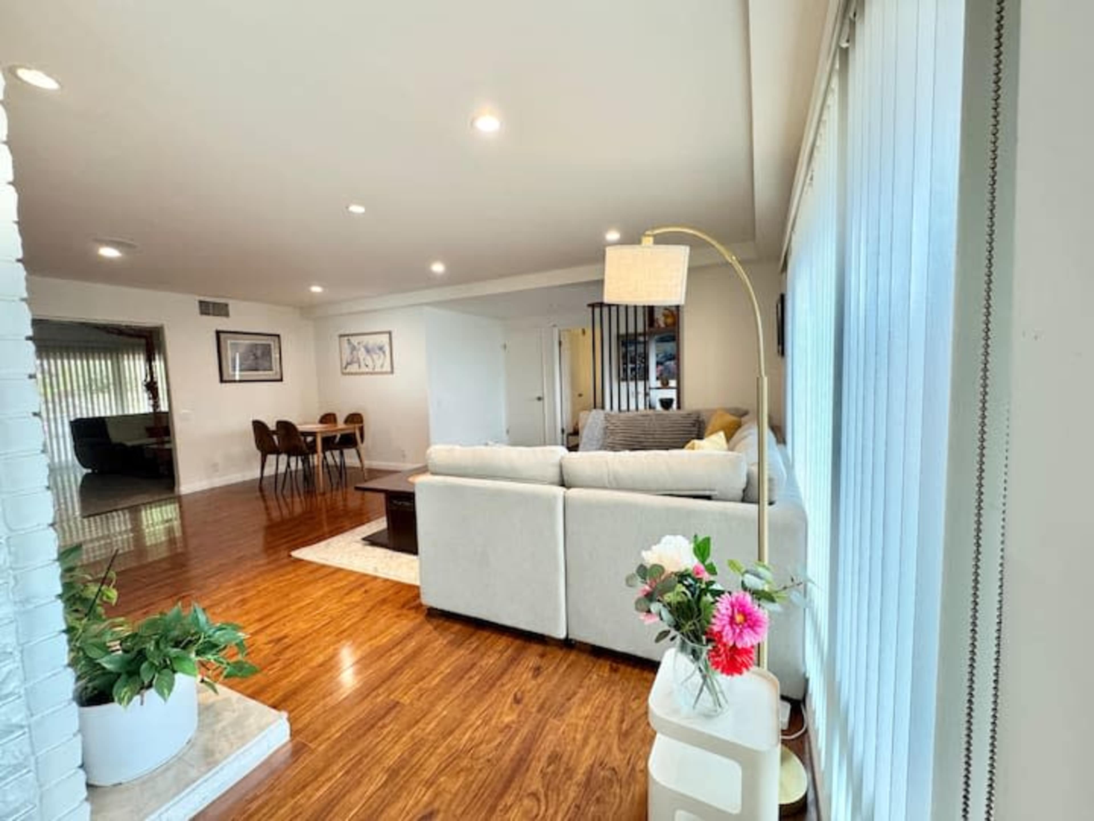 The image shows a bright, open living room featuring a light-colored sectional sofa, wooden flooring, and a view of a dining area in the background.