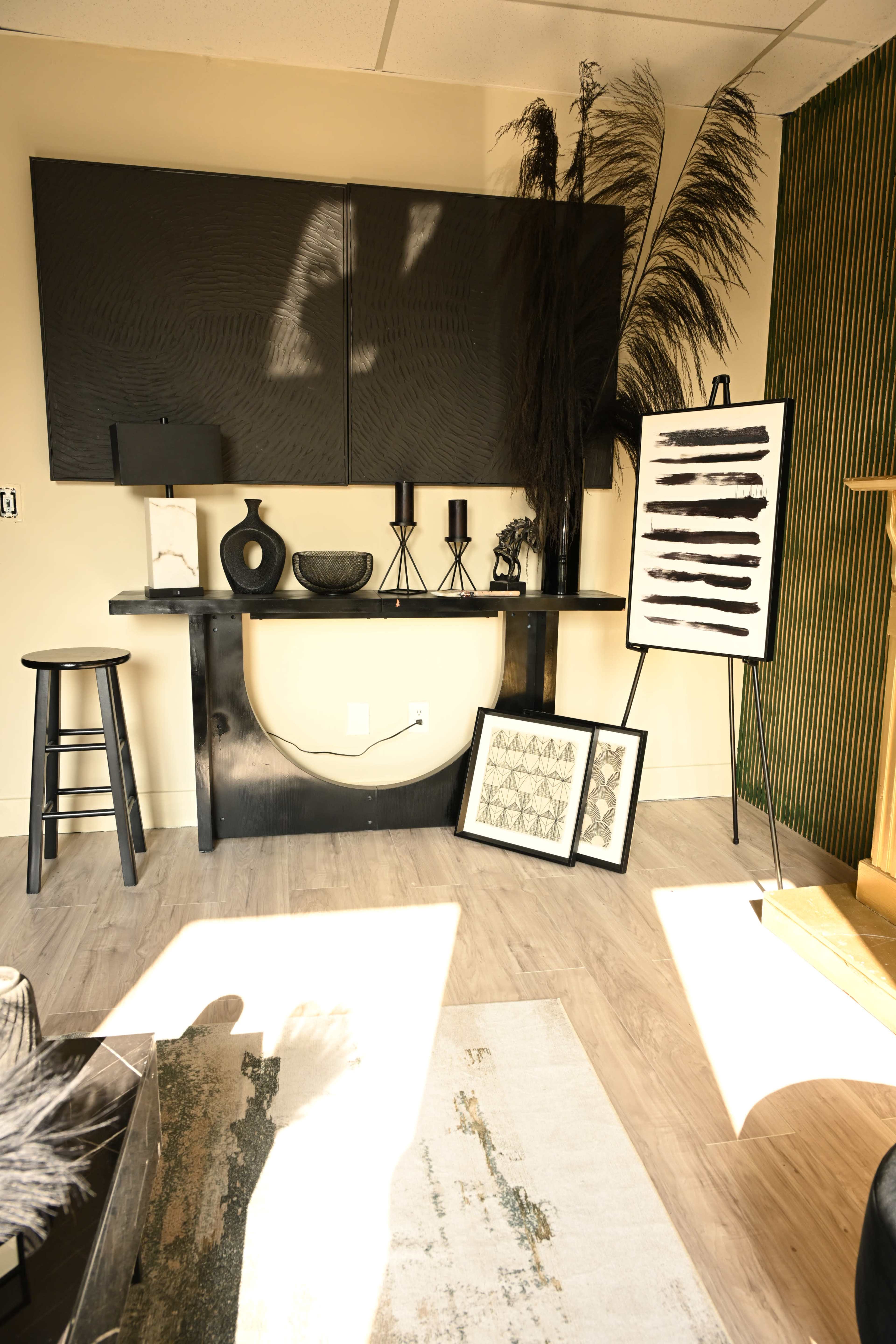 The image shows a modern interior space featuring a black console table with decorative items, an easel displaying artwork, and a striped wall in the background.