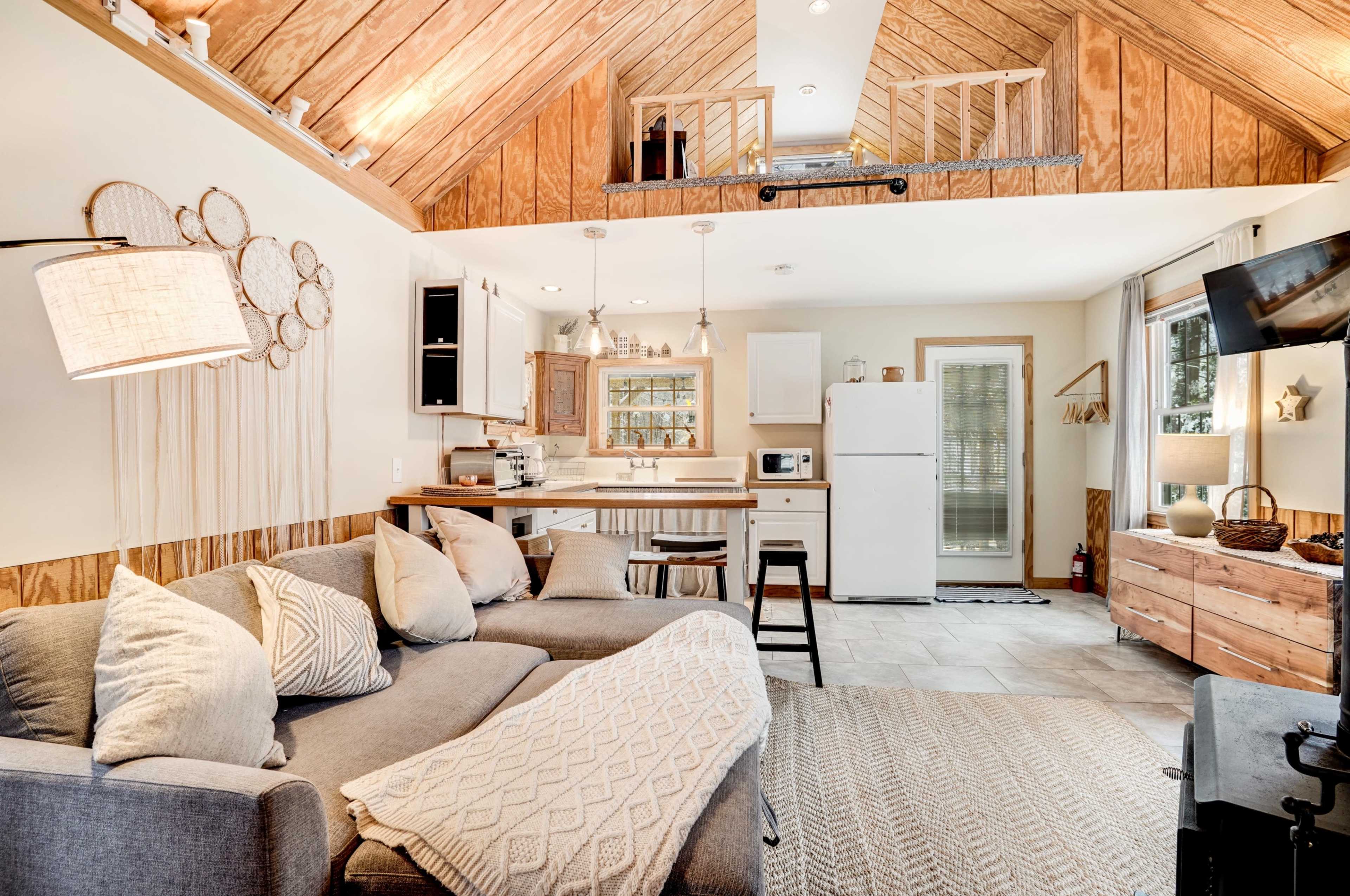 The image shows a cozy living space featuring a gray sofa, a kitchenette with a white refrigerator, and wooden accents on the walls and ceiling.
