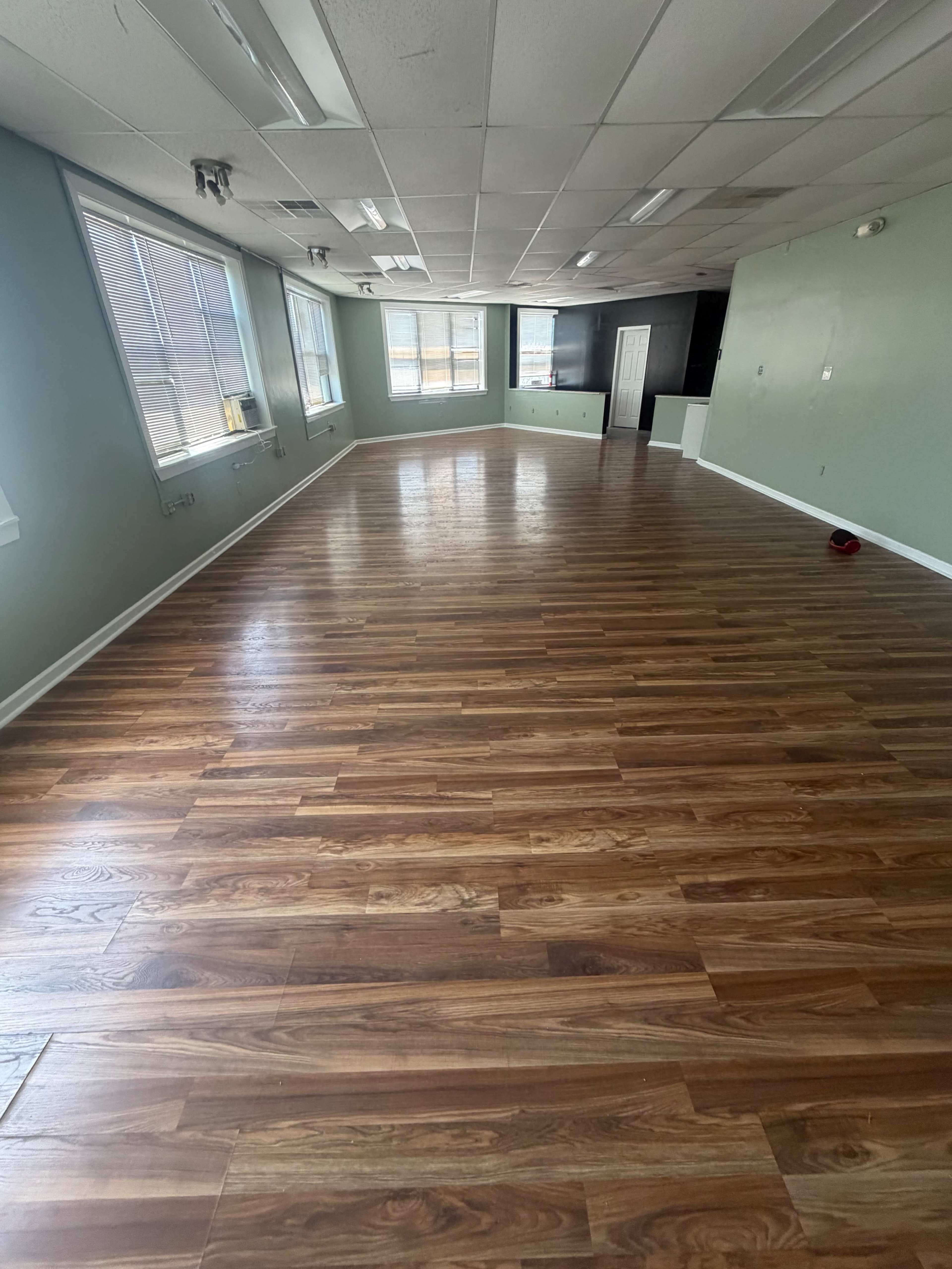 The image shows a spacious, empty room with wooden flooring, large windows, and light green walls.