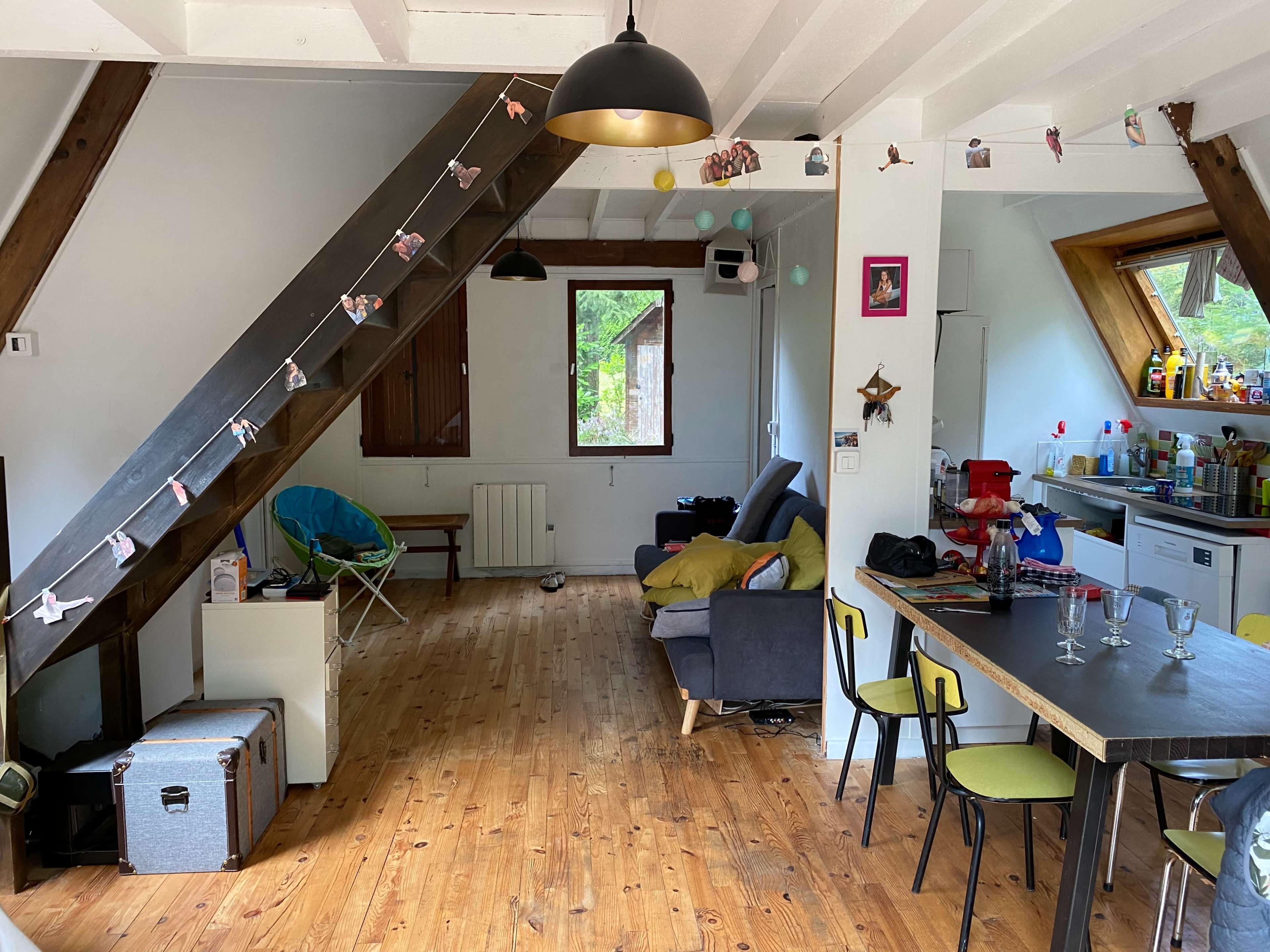 The image shows a cozy living area with wooden flooring, featuring a mix of seating options, a dining table, and a kitchenette in the background, all under a sloped ceiling.