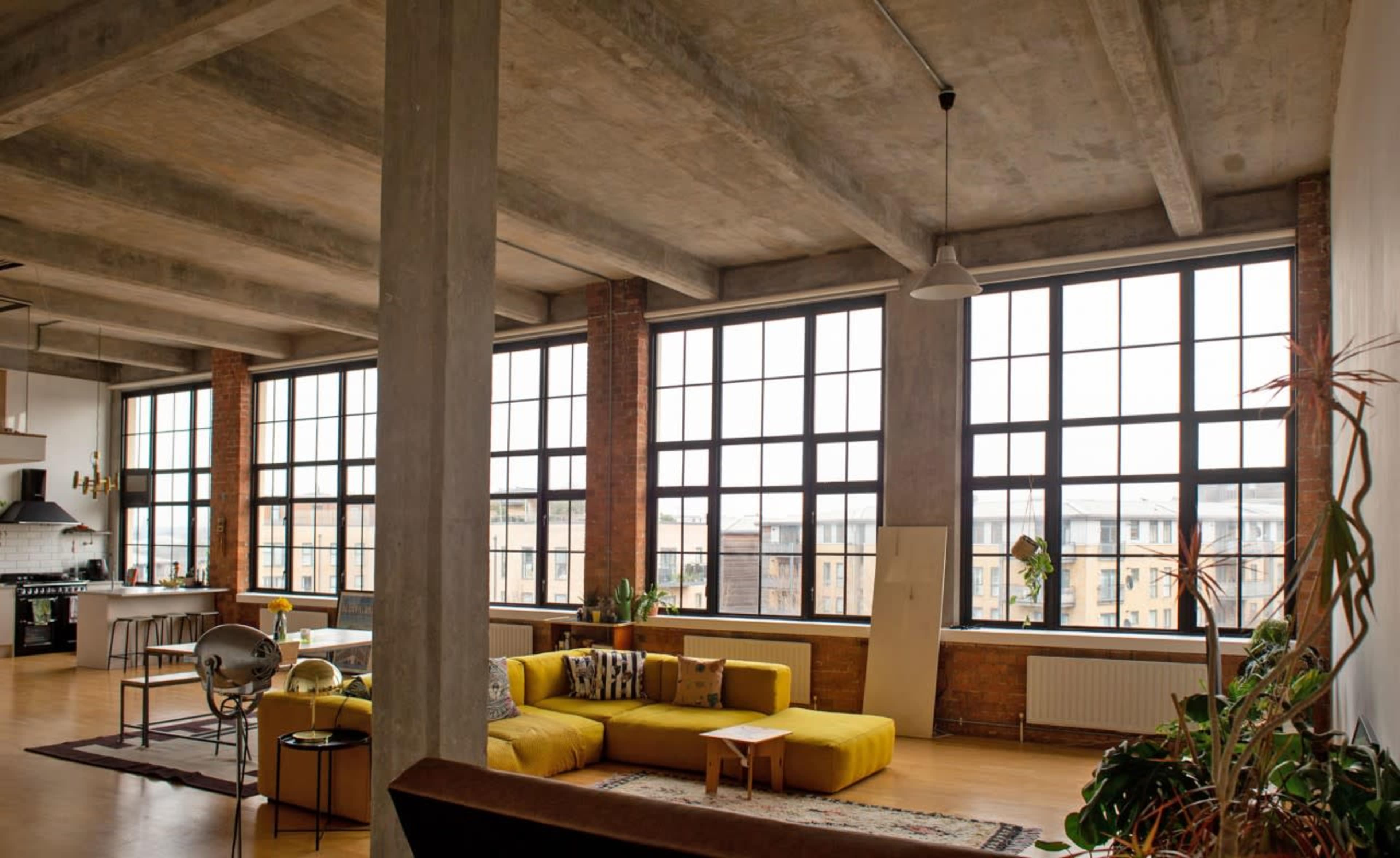 A spacious urban loft with large windows, exposed concrete beams, and casual furniture arranged around a central area.