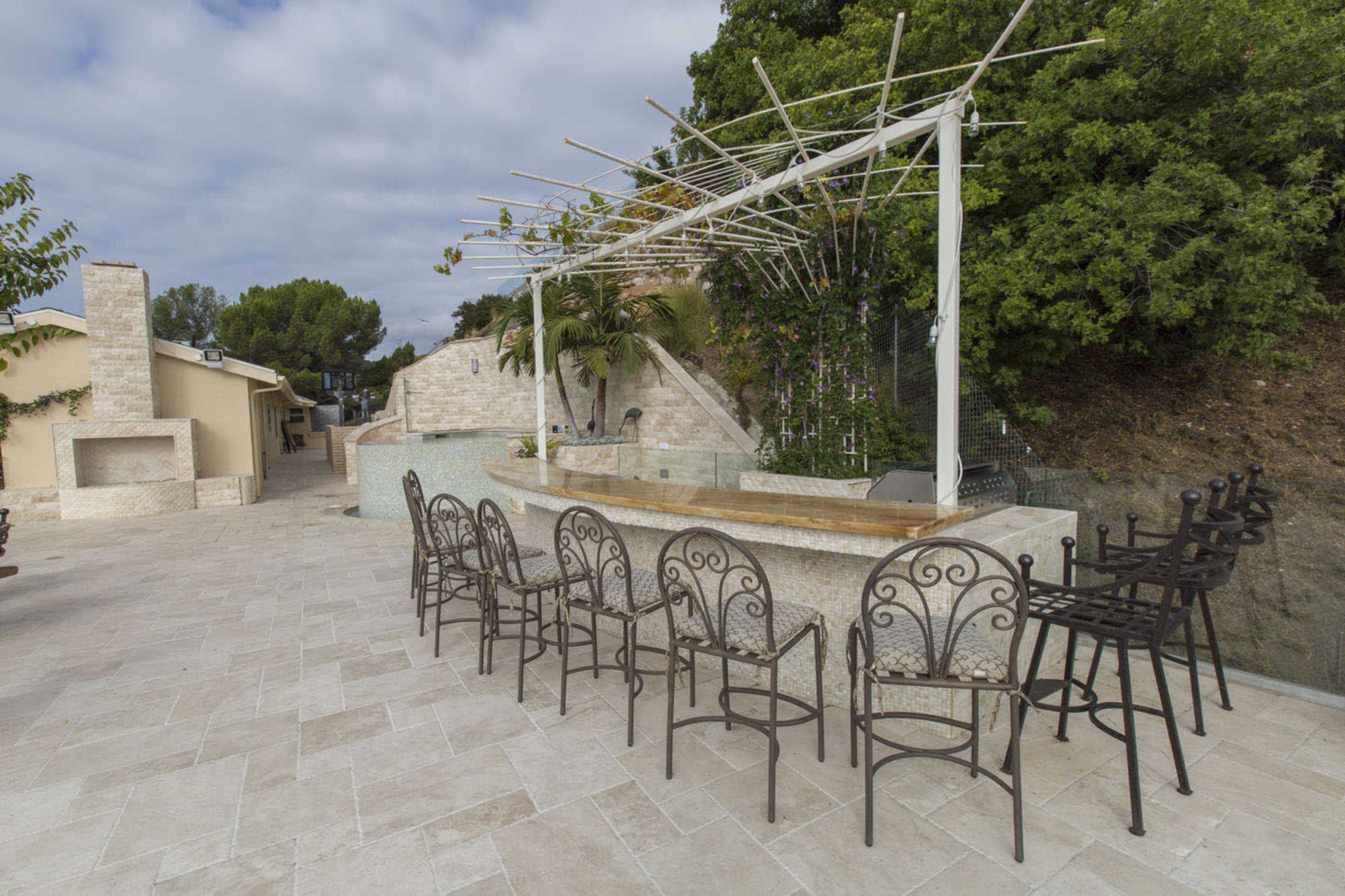 The image shows a bar area with high stools, surrounded by stone pathways and greenery, adjacent to a residential structure.