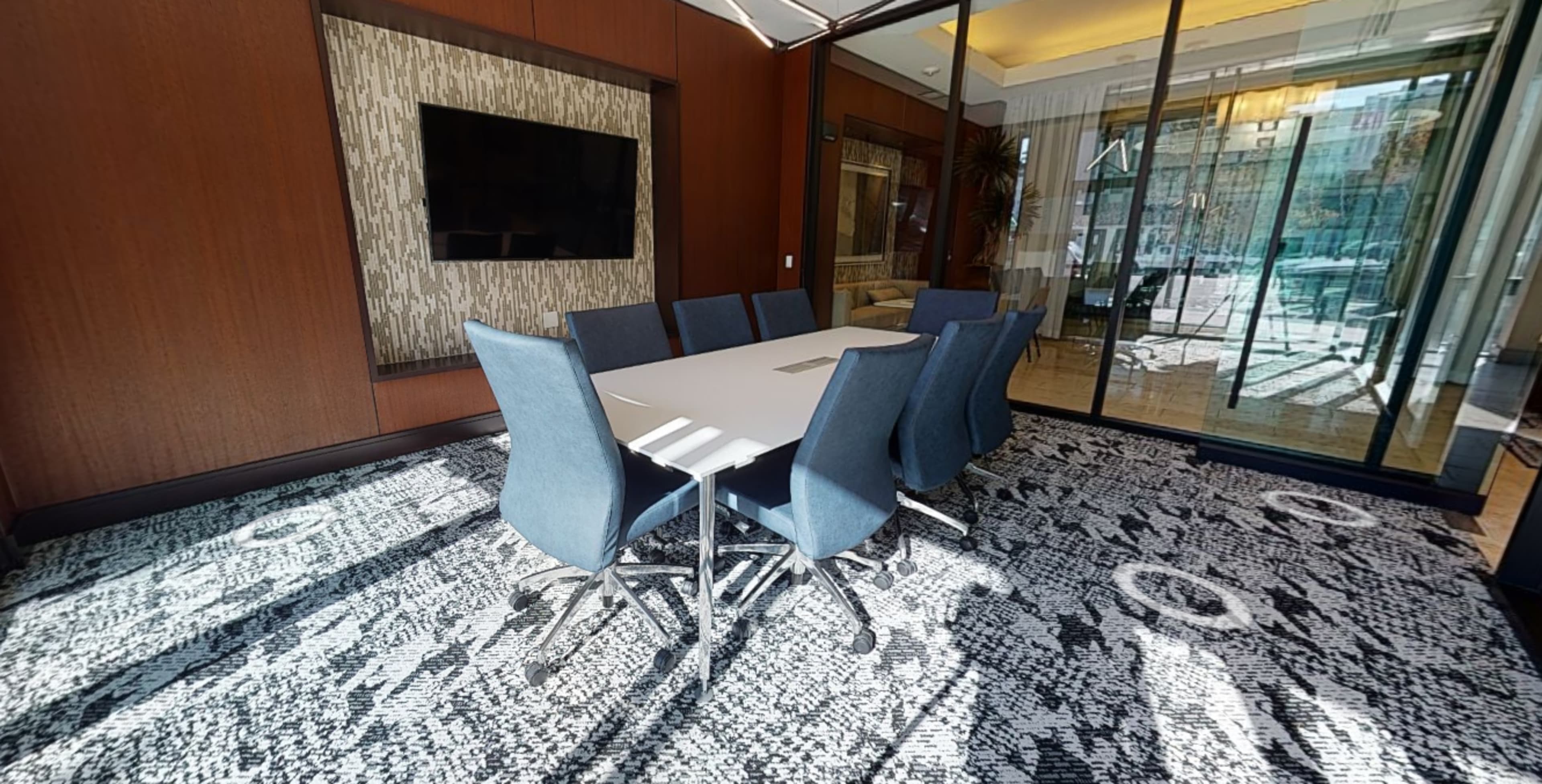 A conference room with a large white table surrounded by eight blue chairs, and glass walls that lead to another room with natural light.
