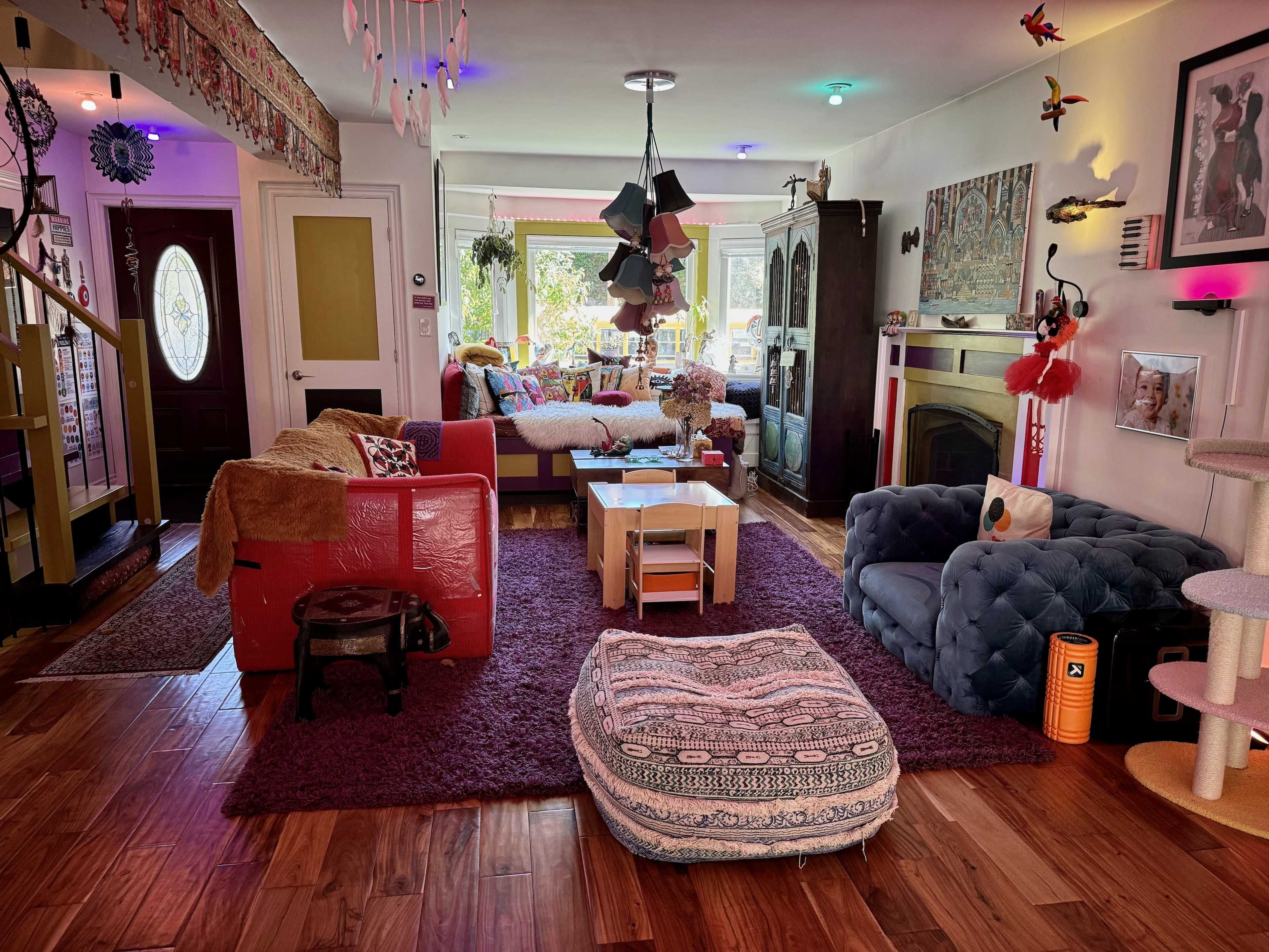 A cozy living room features a mix of colorful furniture, including a red sofa, a blue tufted couch, and a large floor cushion, with an array of decorative items and natural light streaming in through large windows.