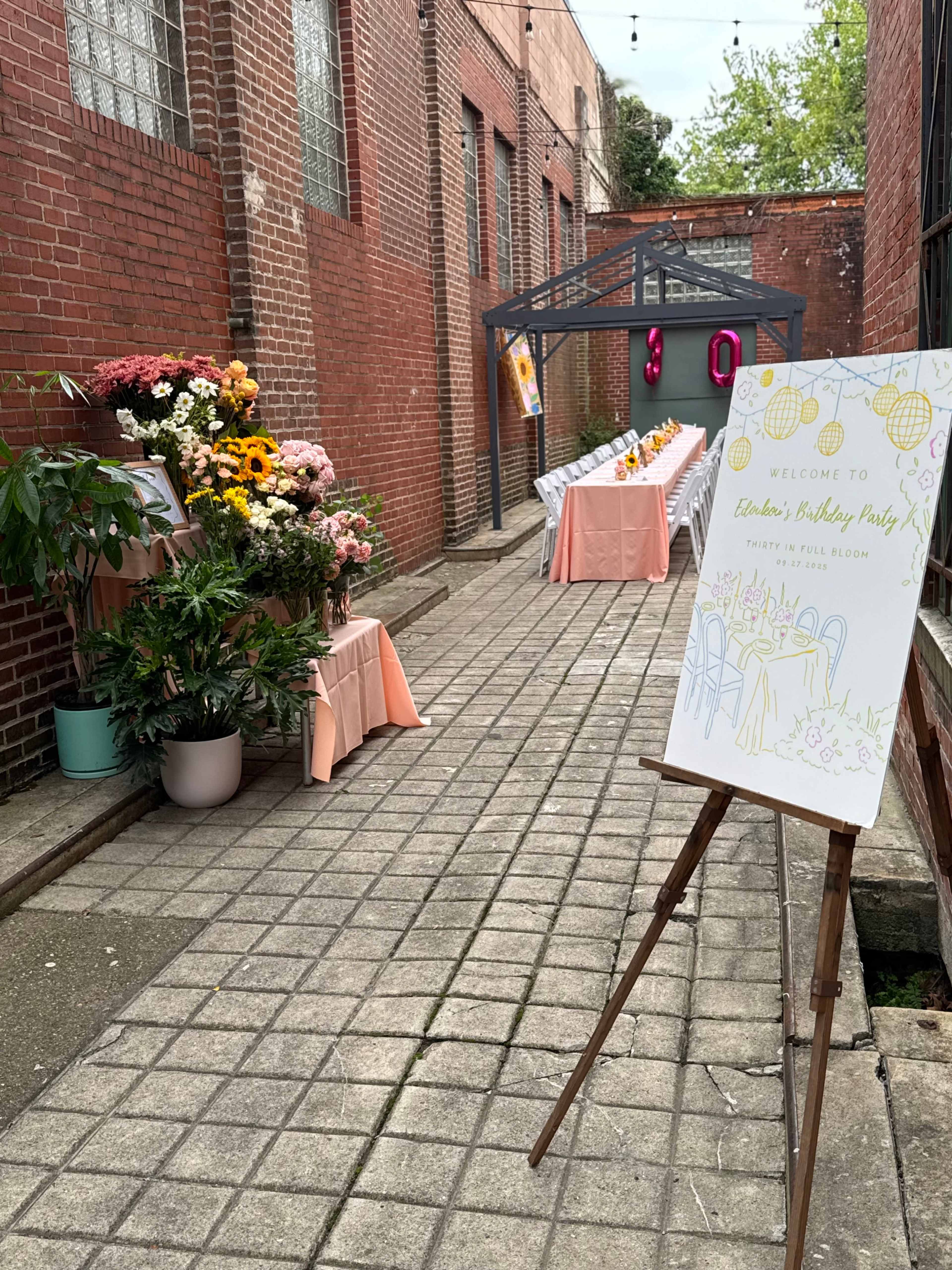The image shows an outdoor venue lined with brick walls, featuring a long table decorated for a birthday party with flowers and a welcome sign.