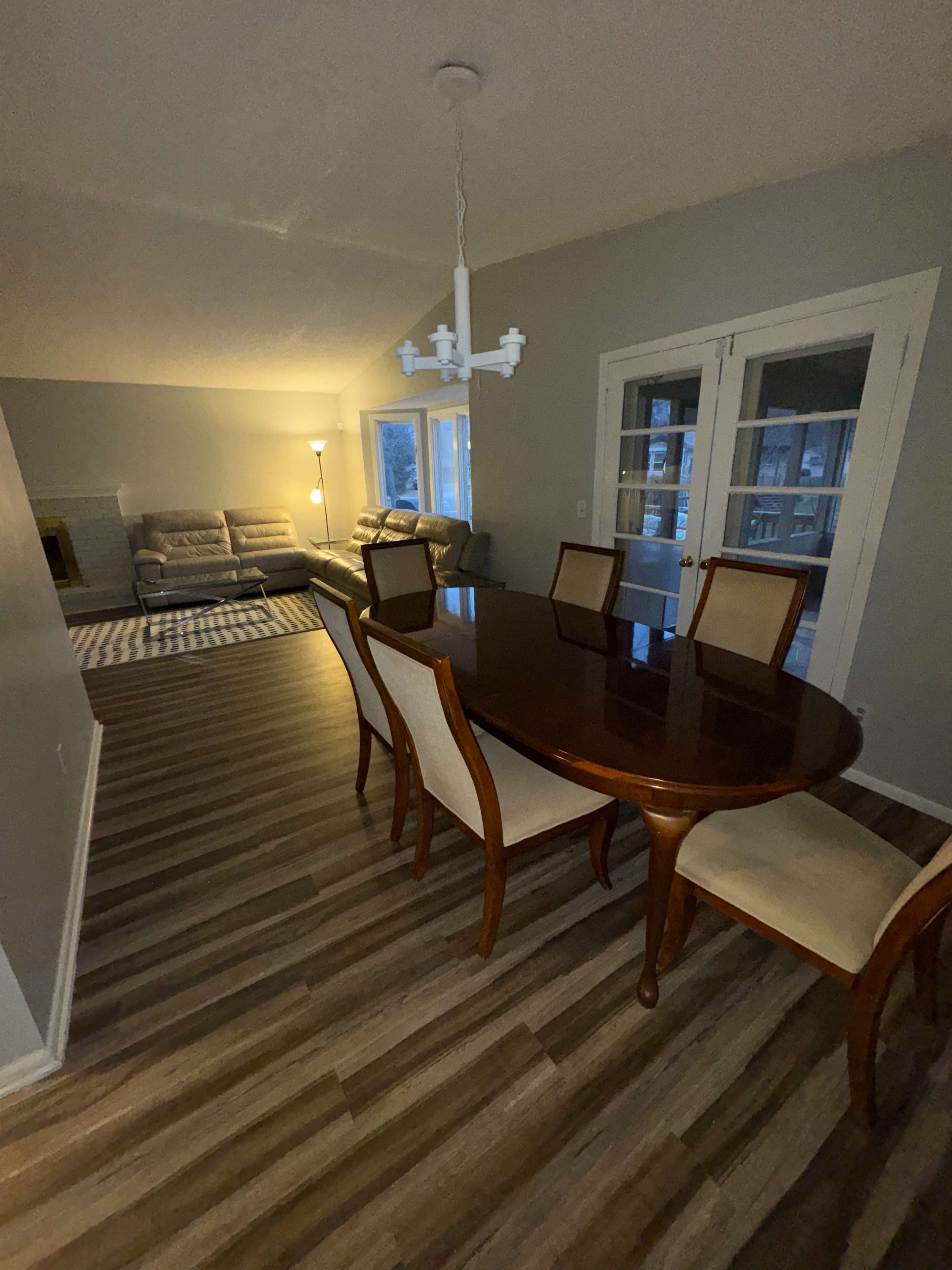 A dining room with a wooden table and chairs, adjacent to a living area that features a couch and a lamp.