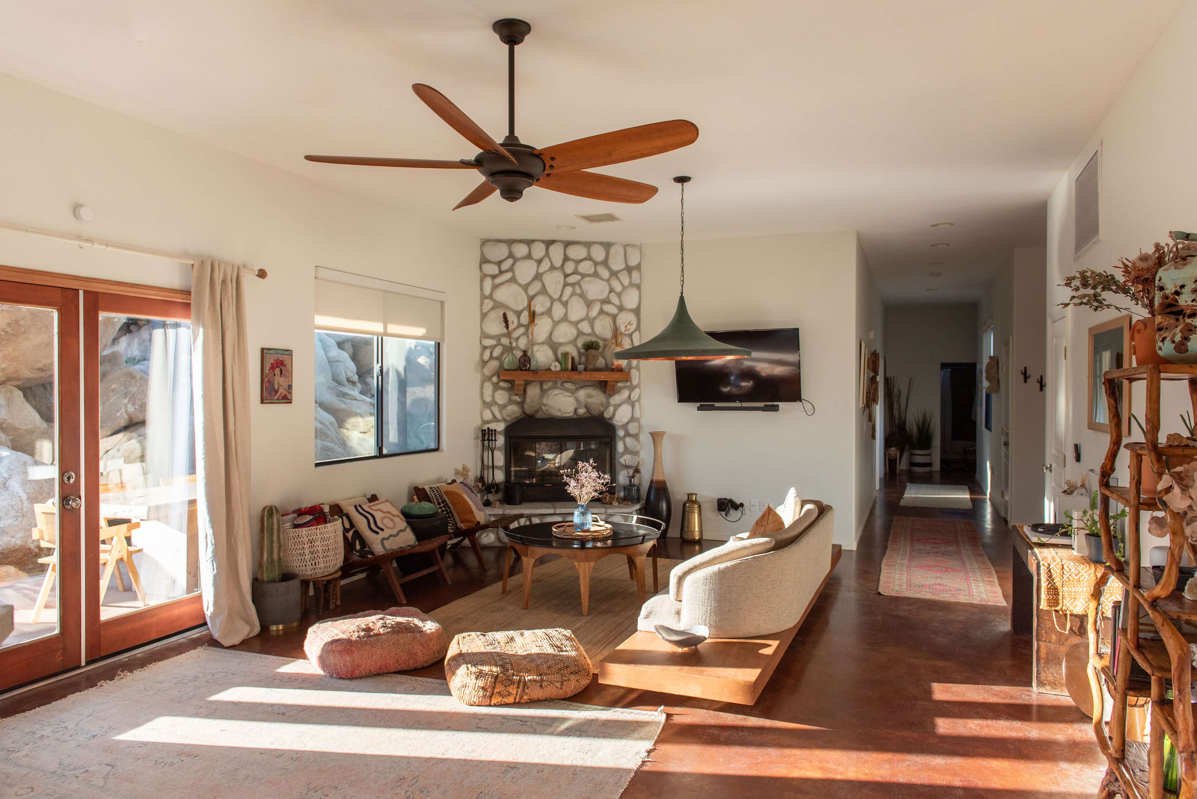 The image shows a cozy living room with a stone fireplace, a ceiling fan, large windows, and a mix of modern and rustic furniture.