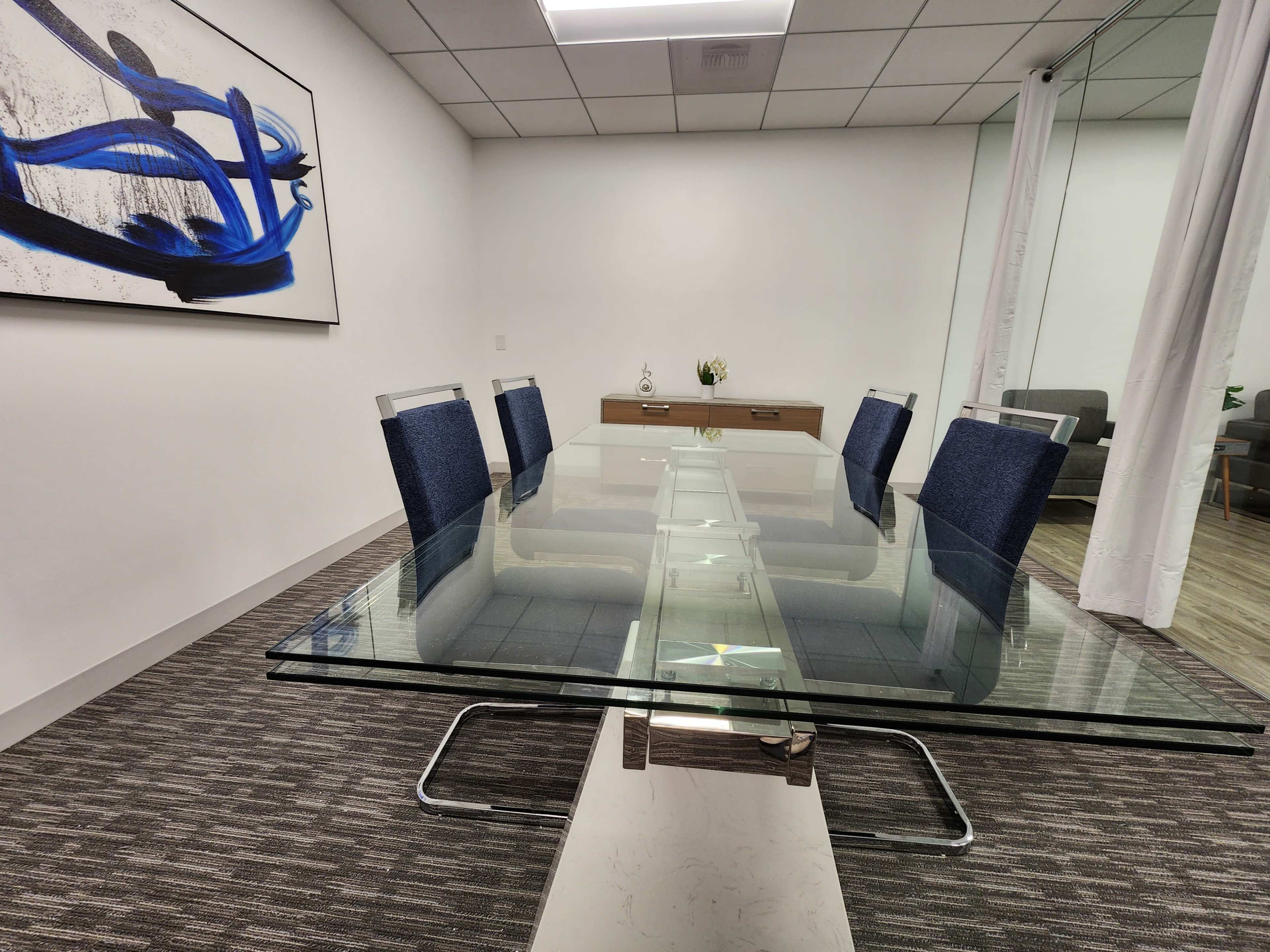A glass conference table with gray carpet is surrounded by six blue chairs in a well-lit meeting room.