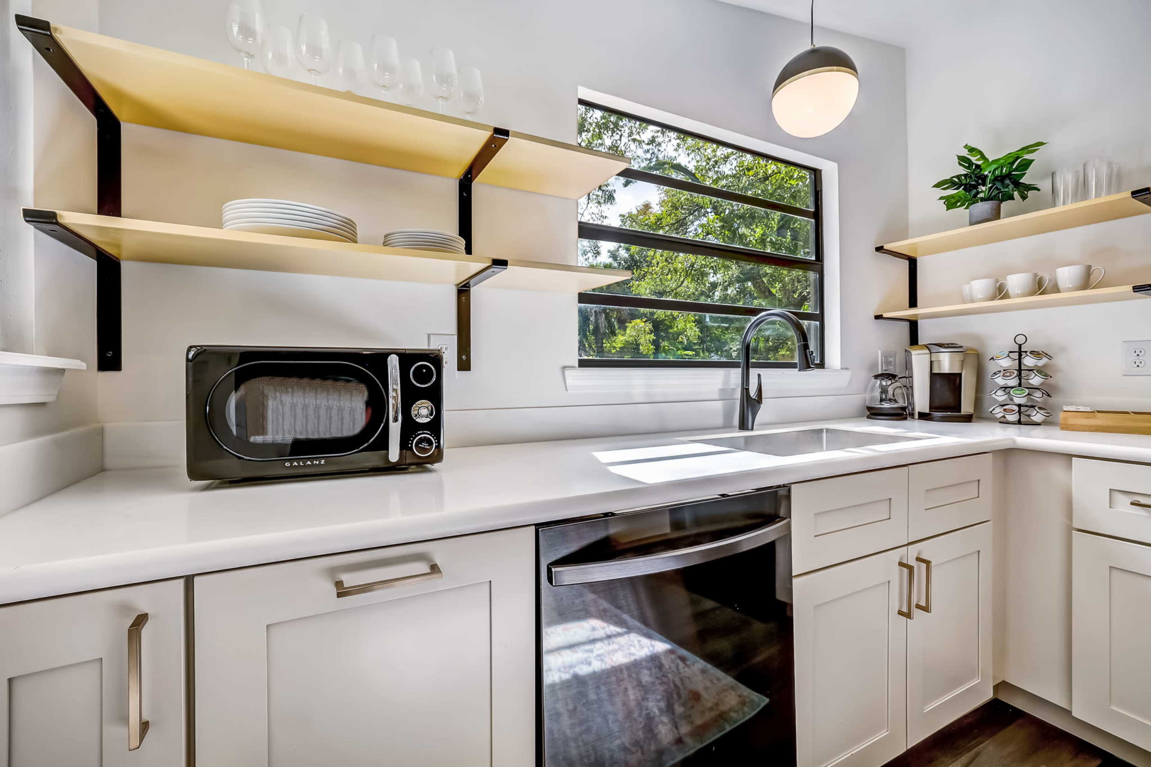 A modern kitchen featuring a stainless steel sink, a microwave, open shelving with plates, and a window allowing natural light to enter.