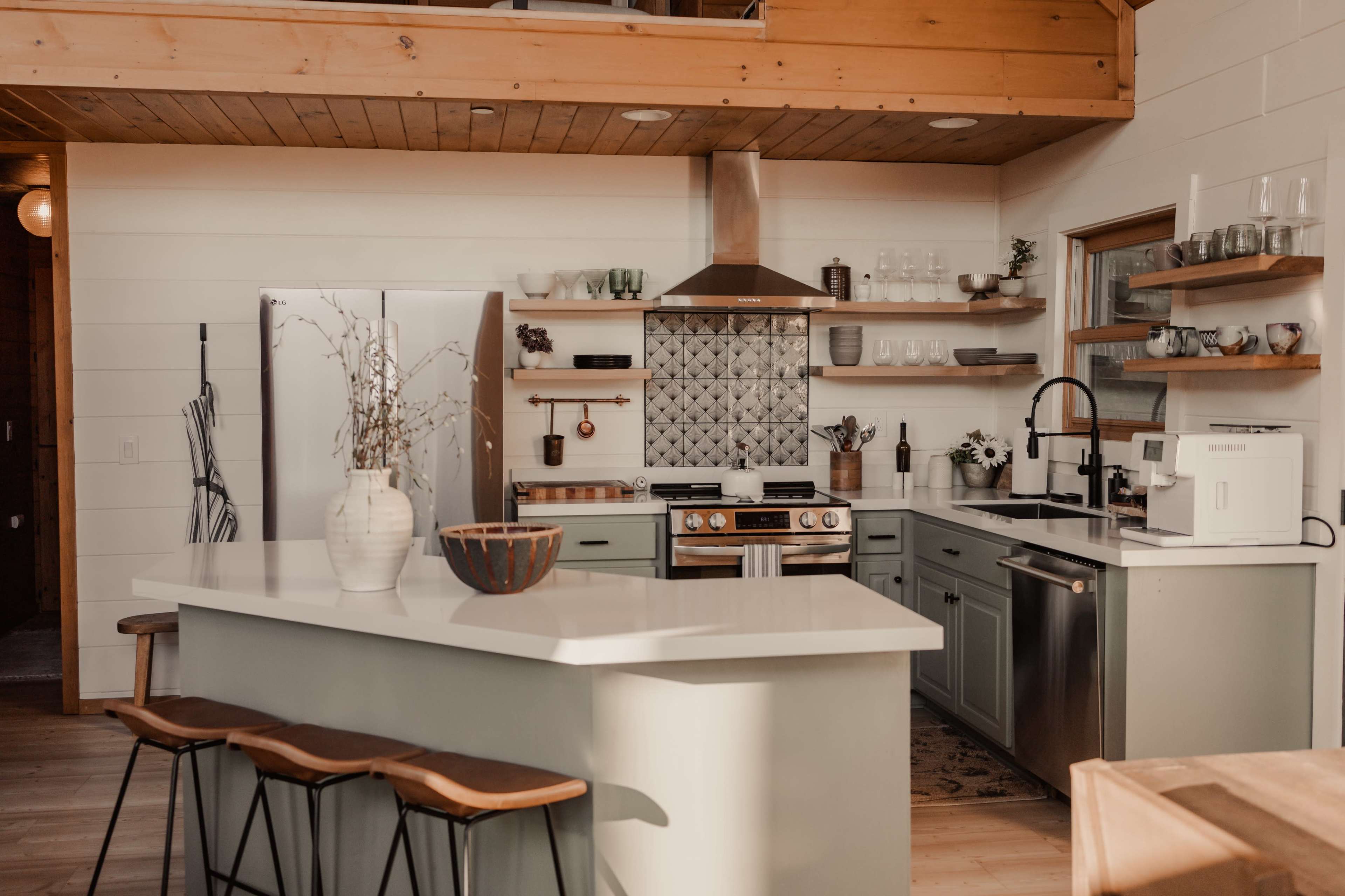 A modern kitchen with a large island, green cabinetry, stainless steel appliances, and decorative shelves.