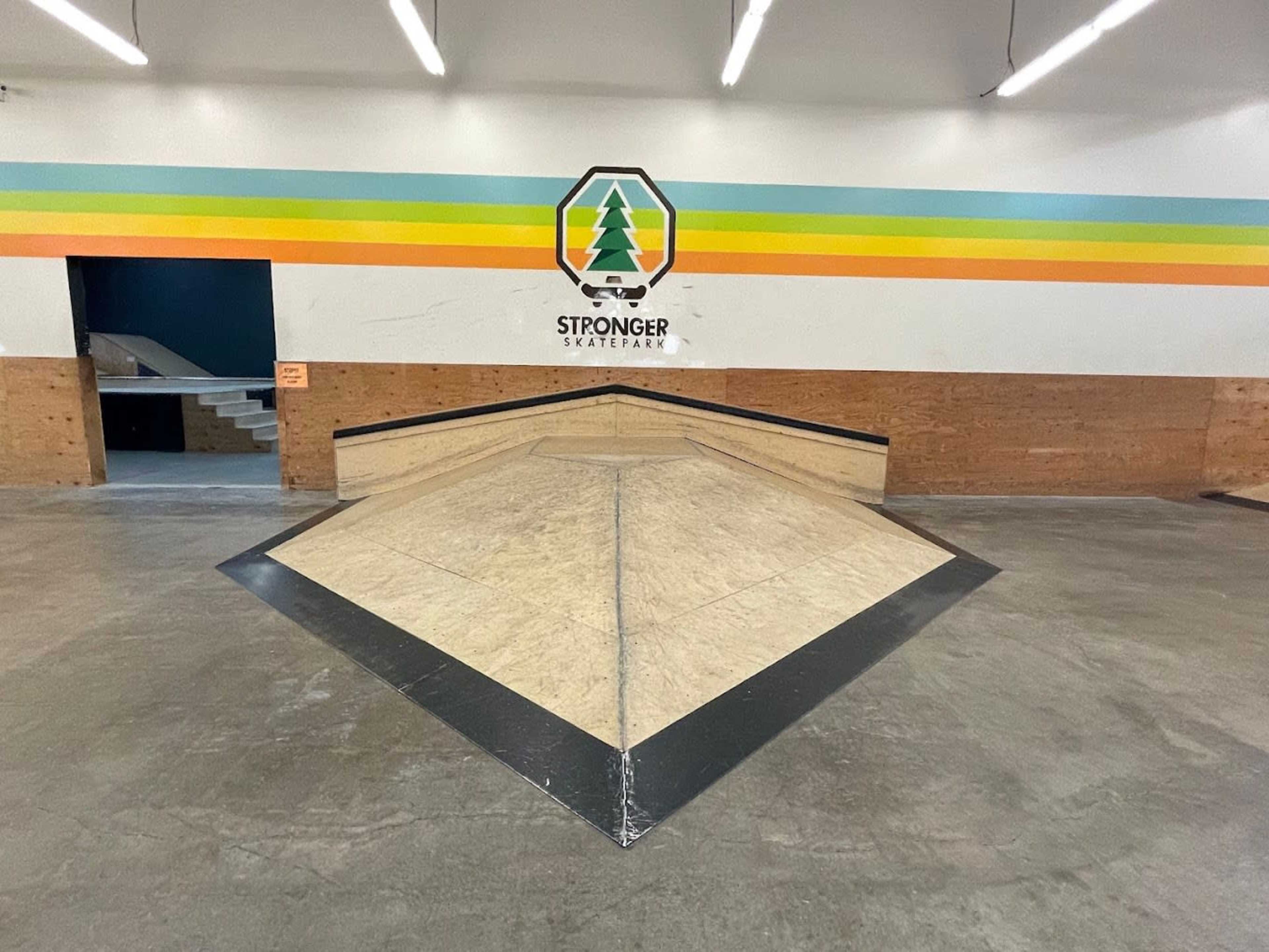 The image shows a skatepark area featuring a large, pointed ramp with wooden surfaces and a colorful striped wall in the background.