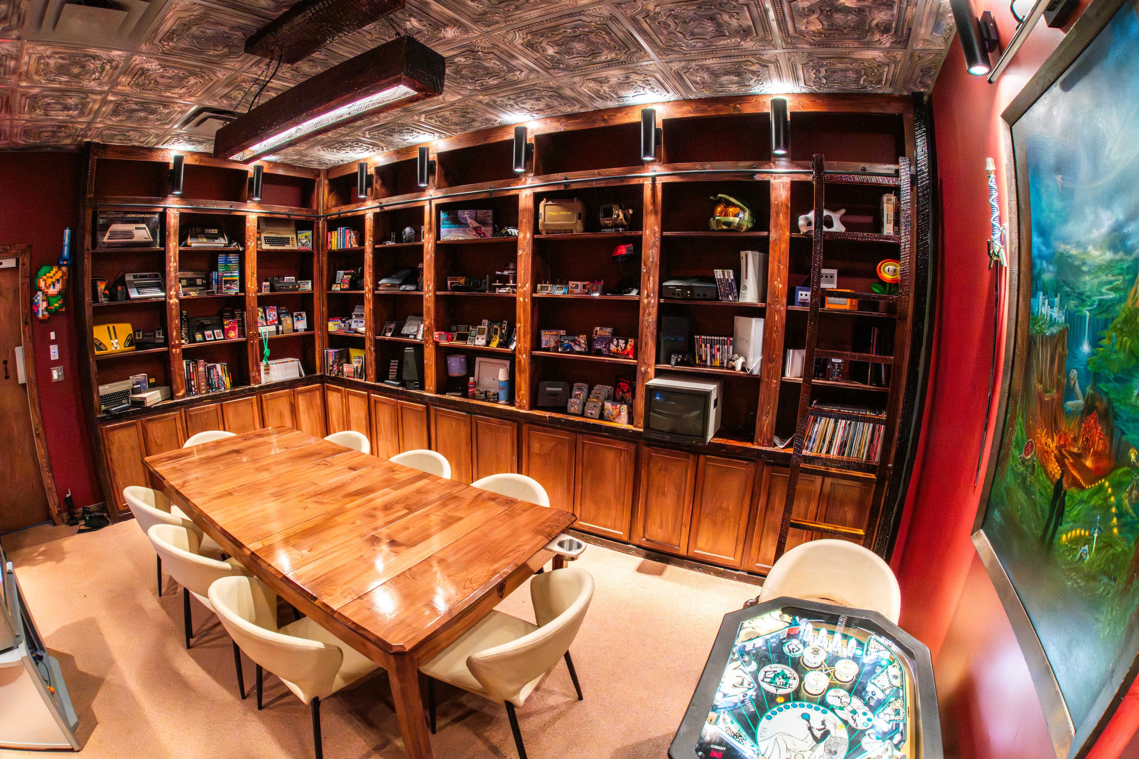 The image shows a room with a large wooden table surrounded by chairs, and a wall unit filled with various gaming consoles and collectibles.