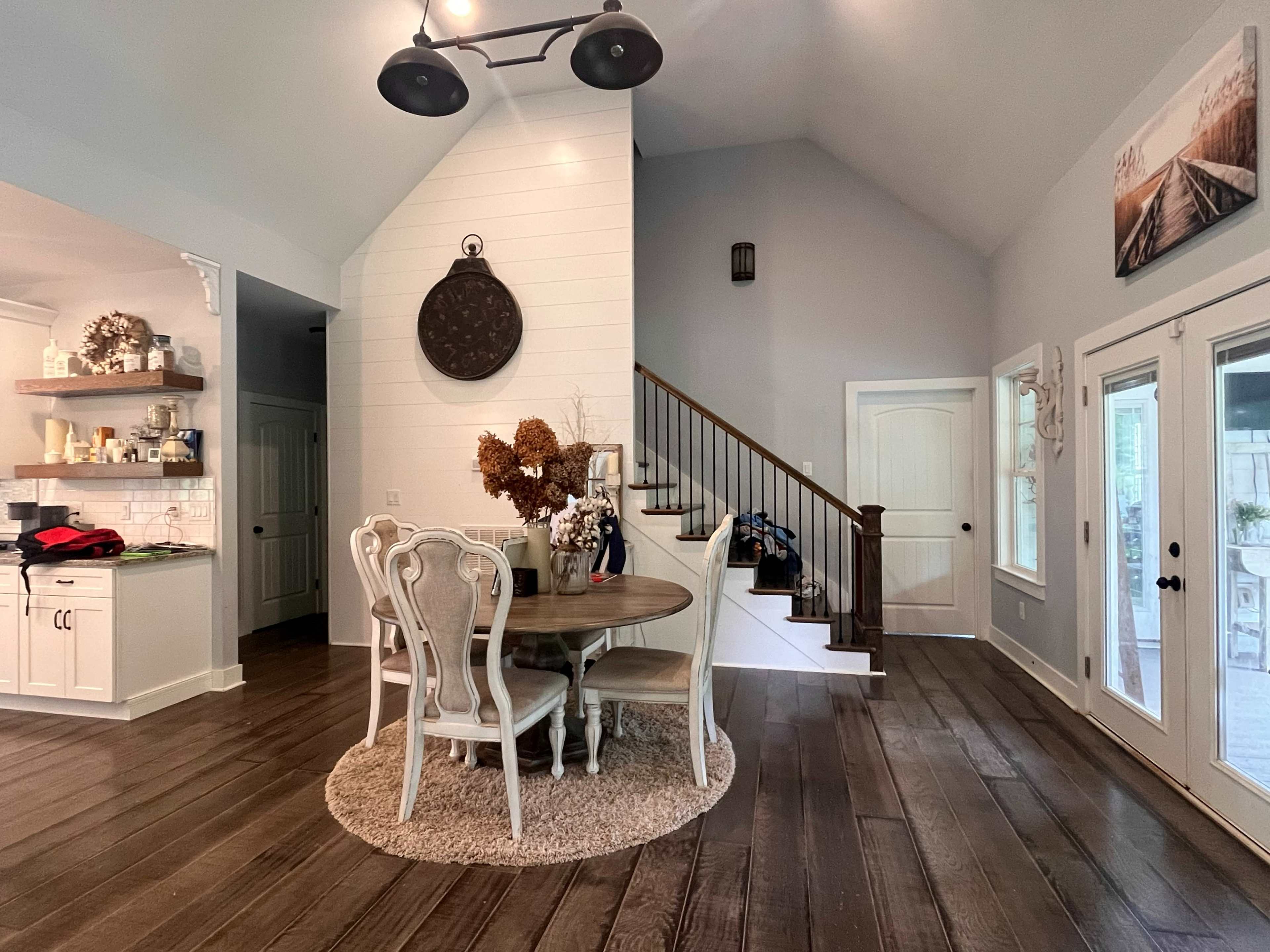 A round dining table with four chairs sits on a circular rug in the open space of a modern home, adjacent to a staircase and kitchen area.