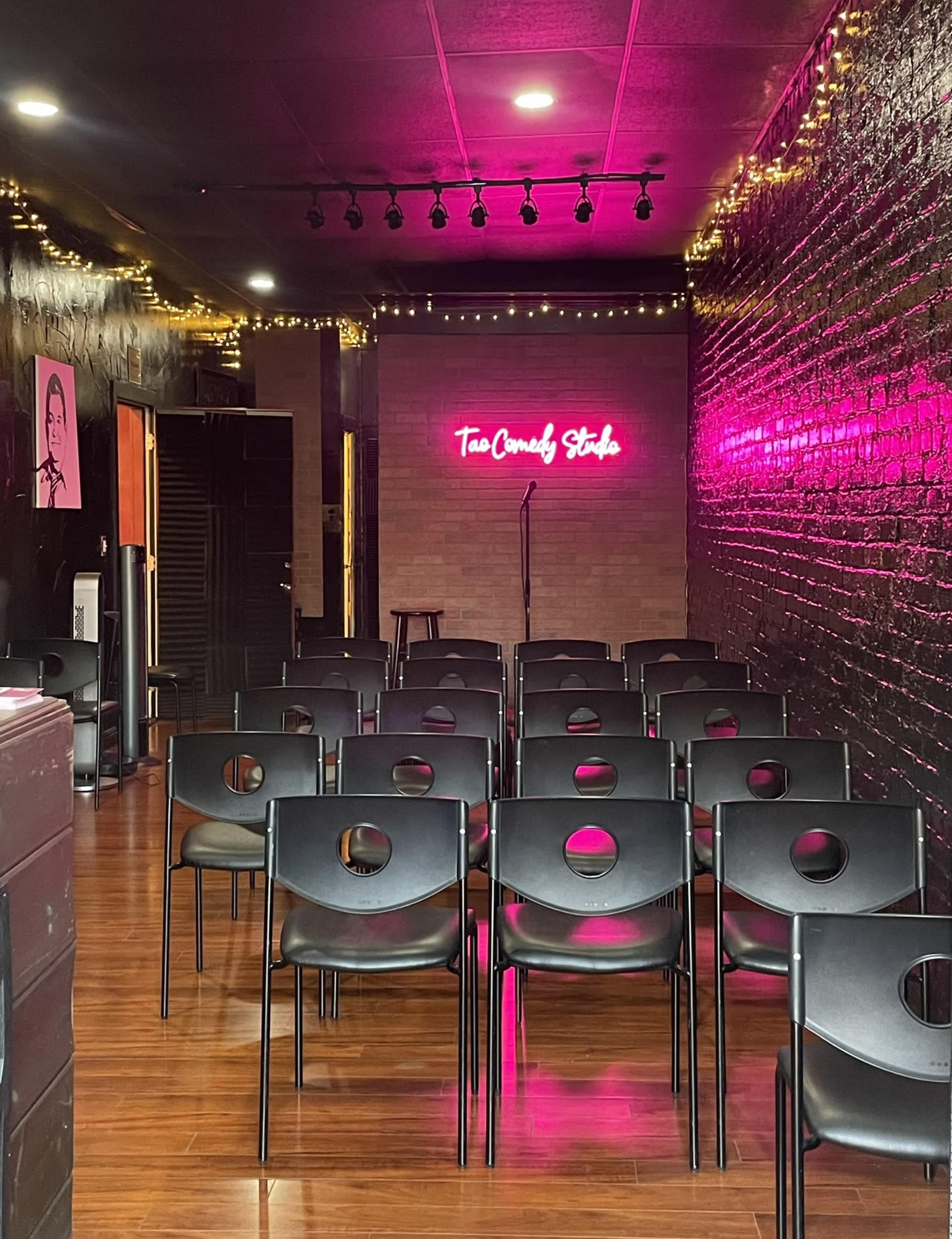 The image shows a small comedy venue with rows of black chairs facing a stage, which has a neon sign that reads "The Comedy Stole."