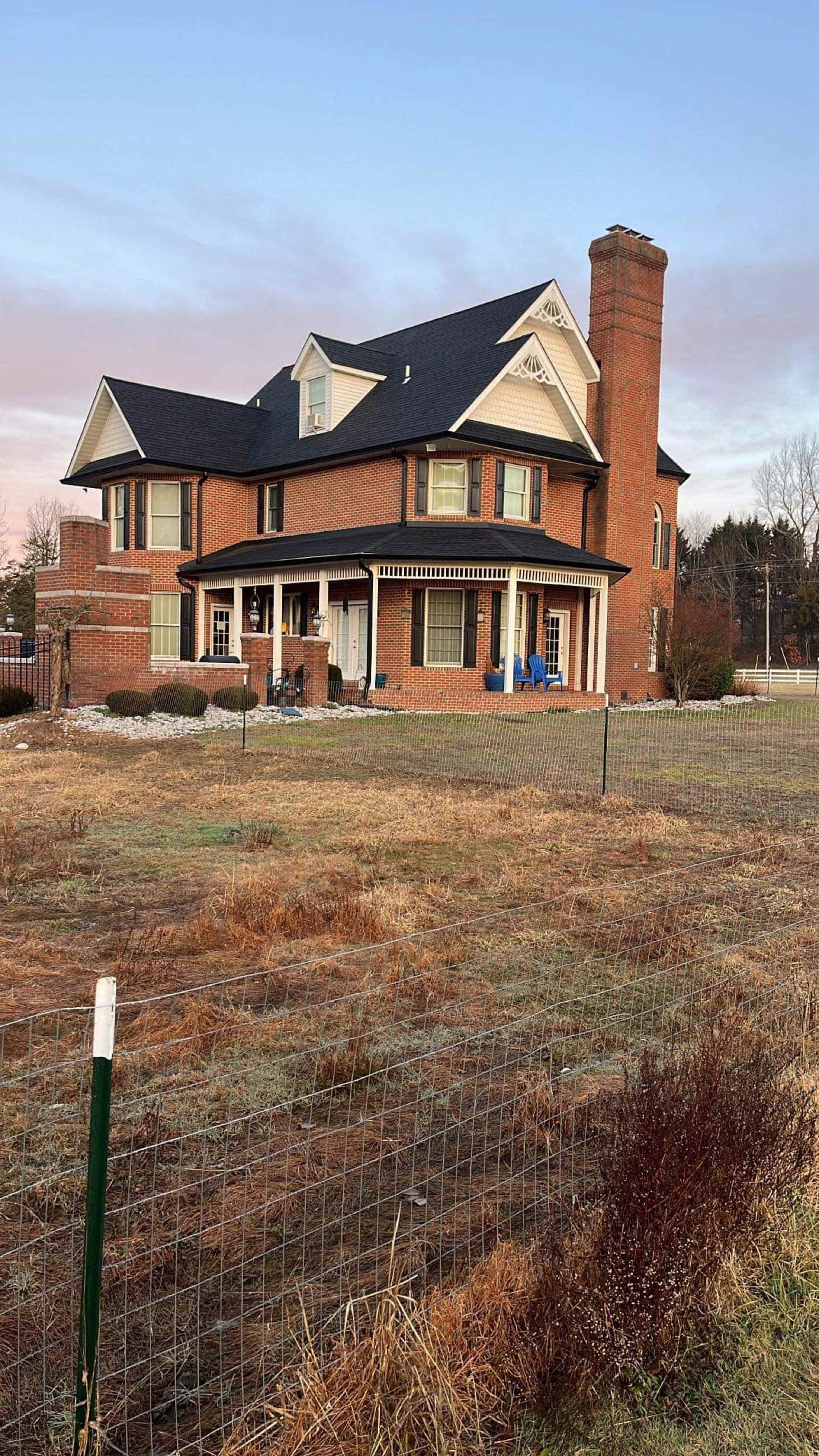 A large, two-story brick house with a peaked roof and a chimney sits on a grassy lot, surrounded by a wooden fence.