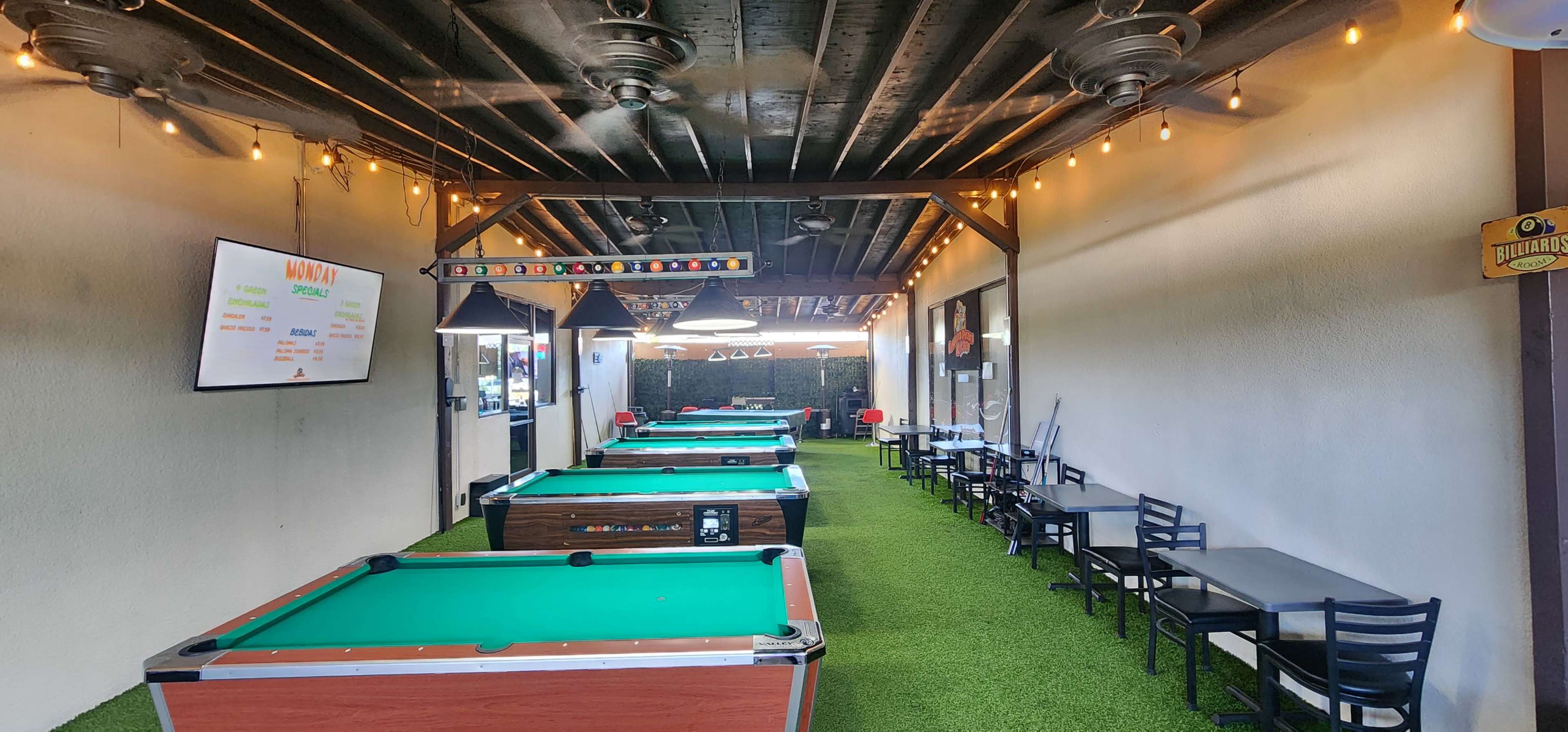 The image shows a well-lit outdoor area featuring several pool tables and tables with chairs on a green carpet, with decorative string lights above.