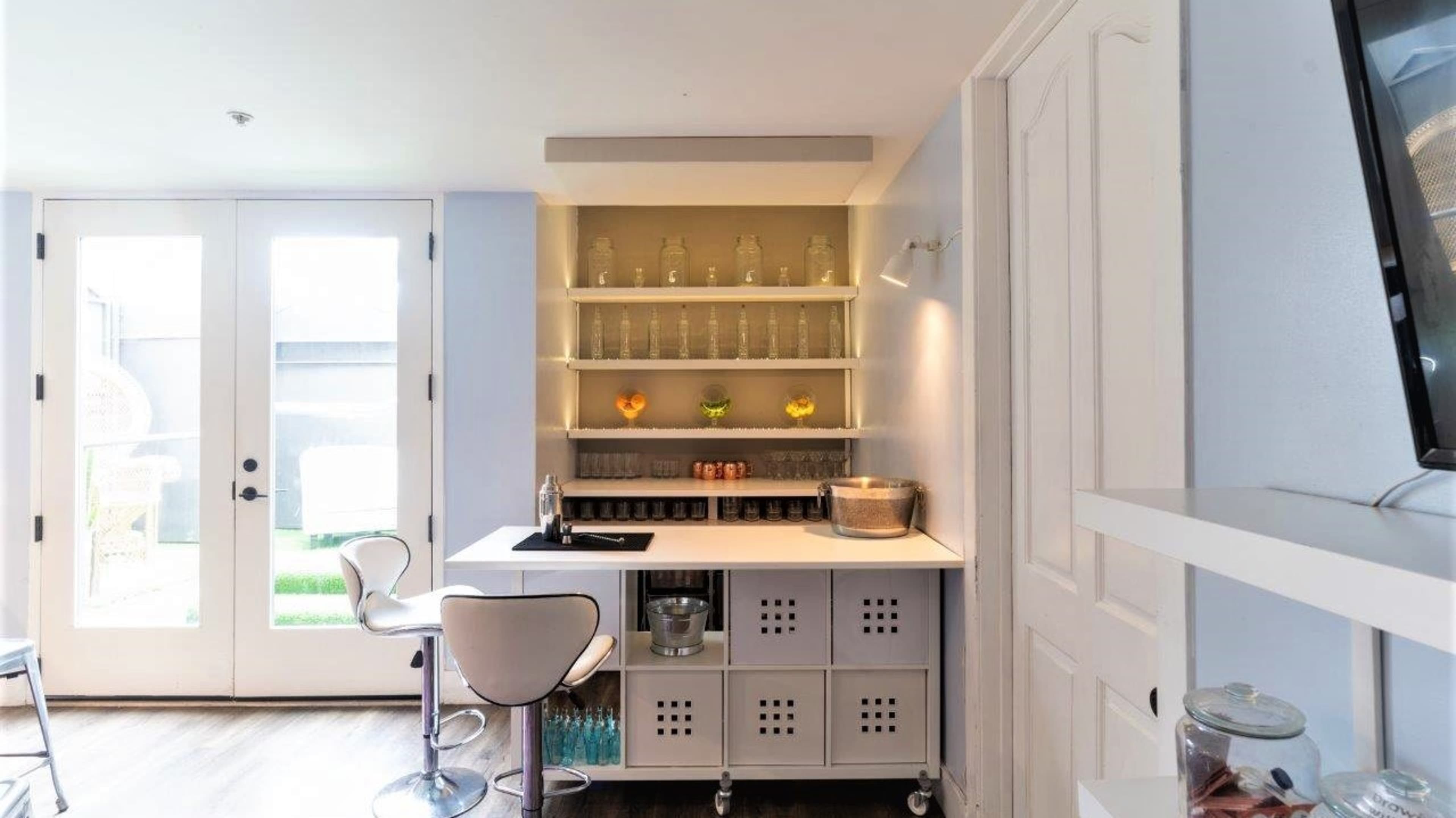 The image shows a modern kitchenette with open shelving displaying glass containers and a small bar area equipped with a sink and seating, beside a set of double doors leading to an outdoor space.