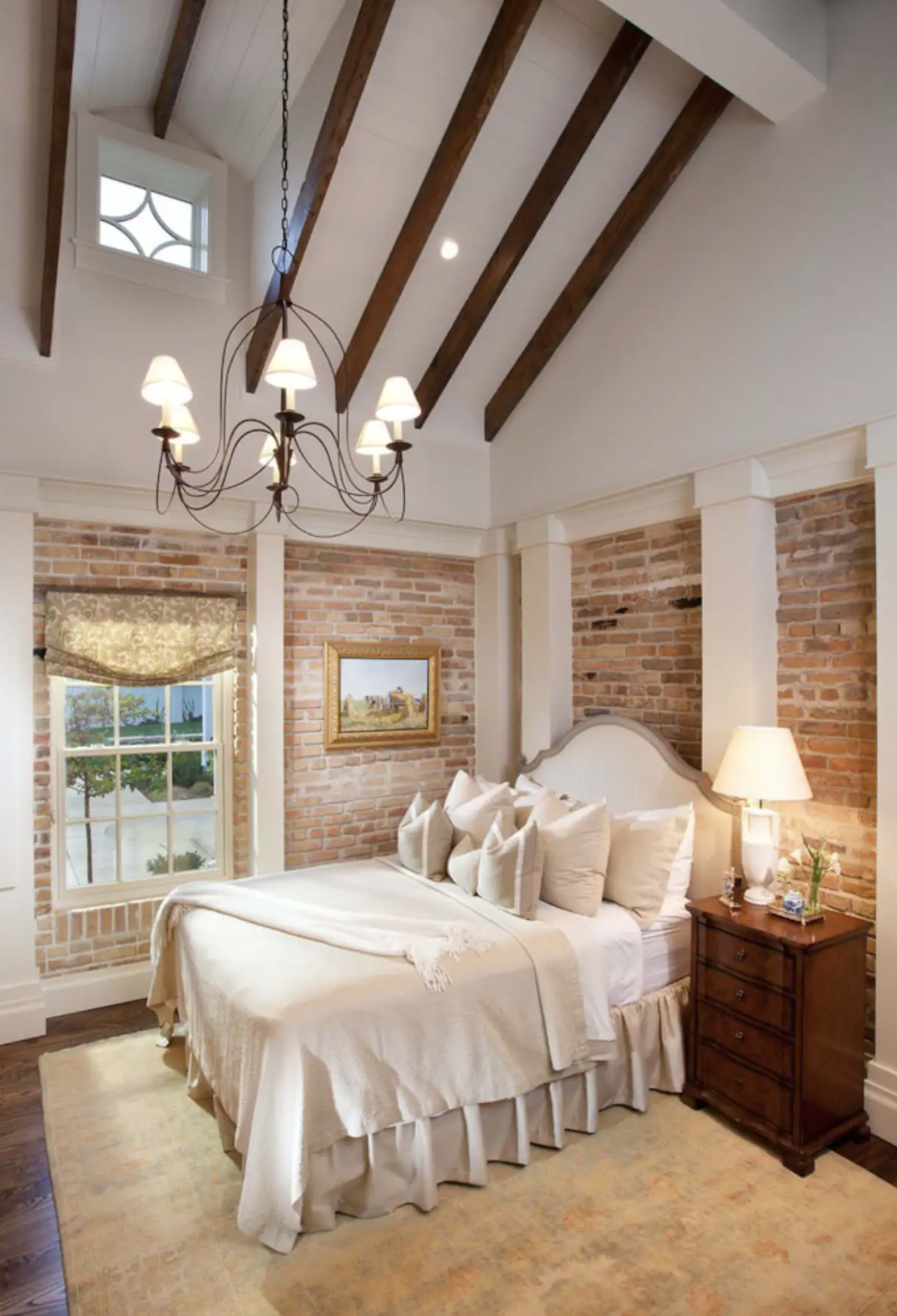 A cozy bedroom featuring a large bed with a light-colored bedspread, brick walls, and a chandelier hanging from the ceiling.