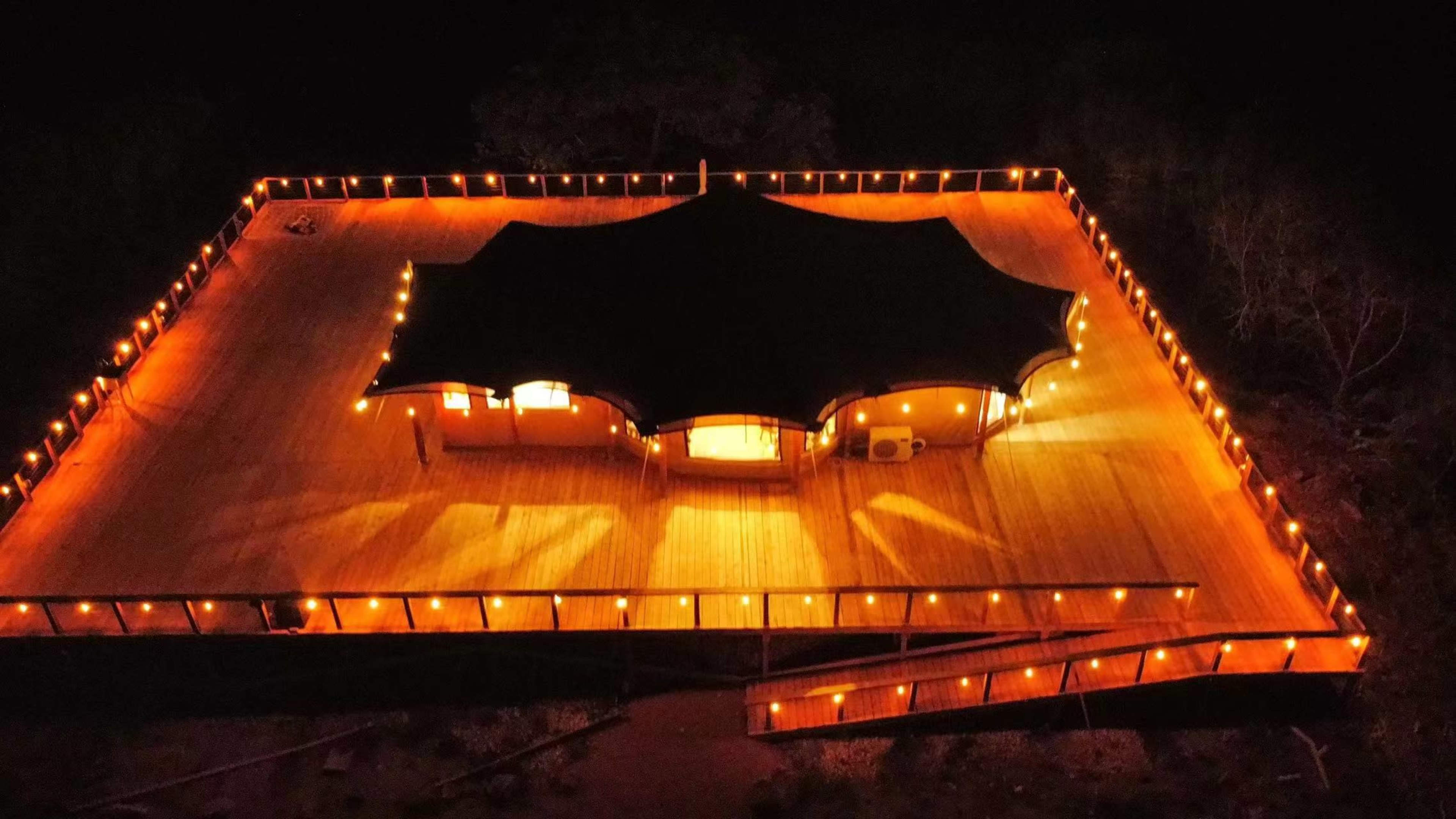 A well-lit, spacious wooden deck surrounds a central tented structure, illuminated by string lights against the nighttime backdrop.