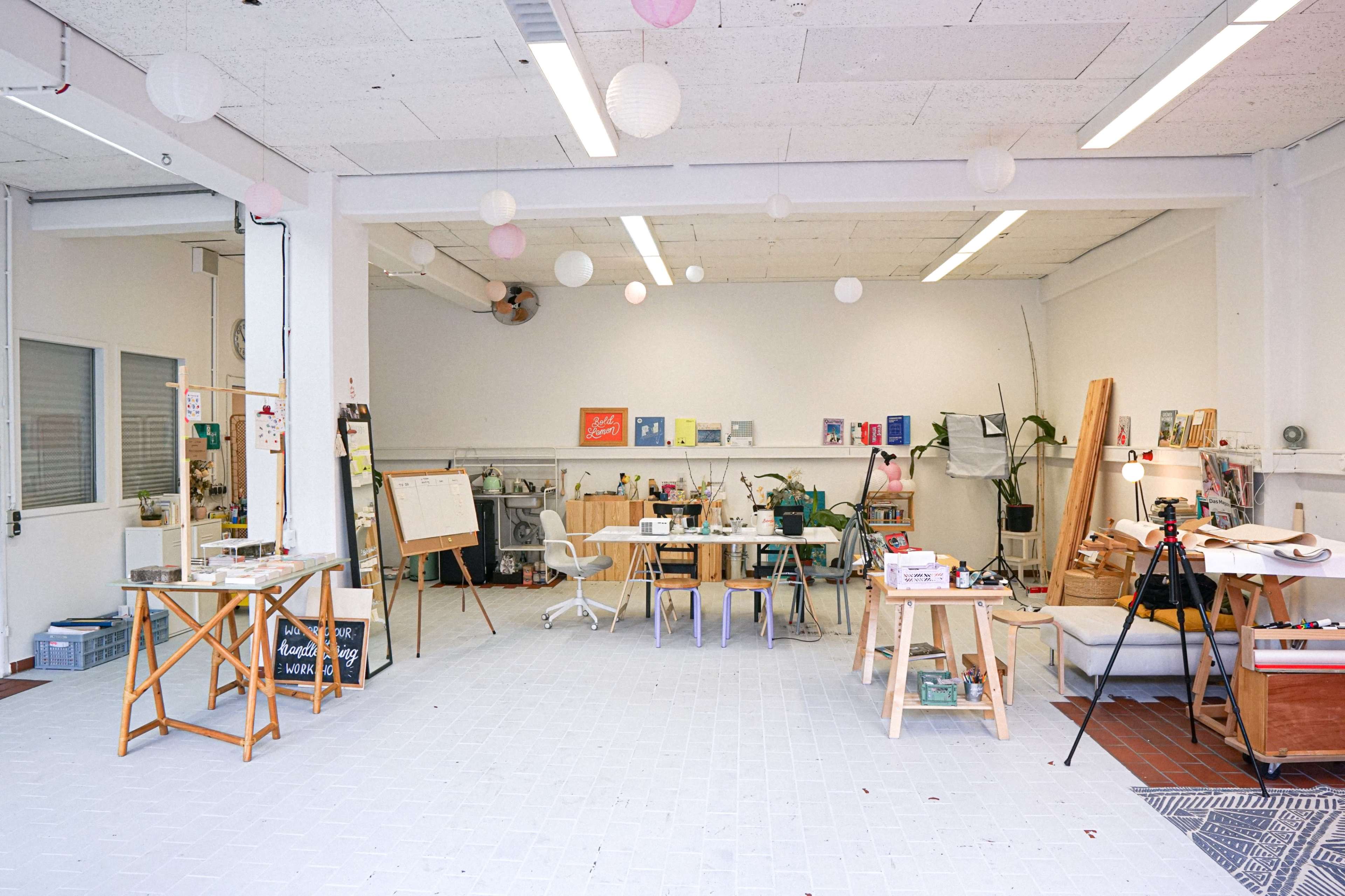 The image shows a spacious, well-lit studio with tables, chairs, and various art supplies scattered around, along with a few decorative lanterns hanging from the ceiling.