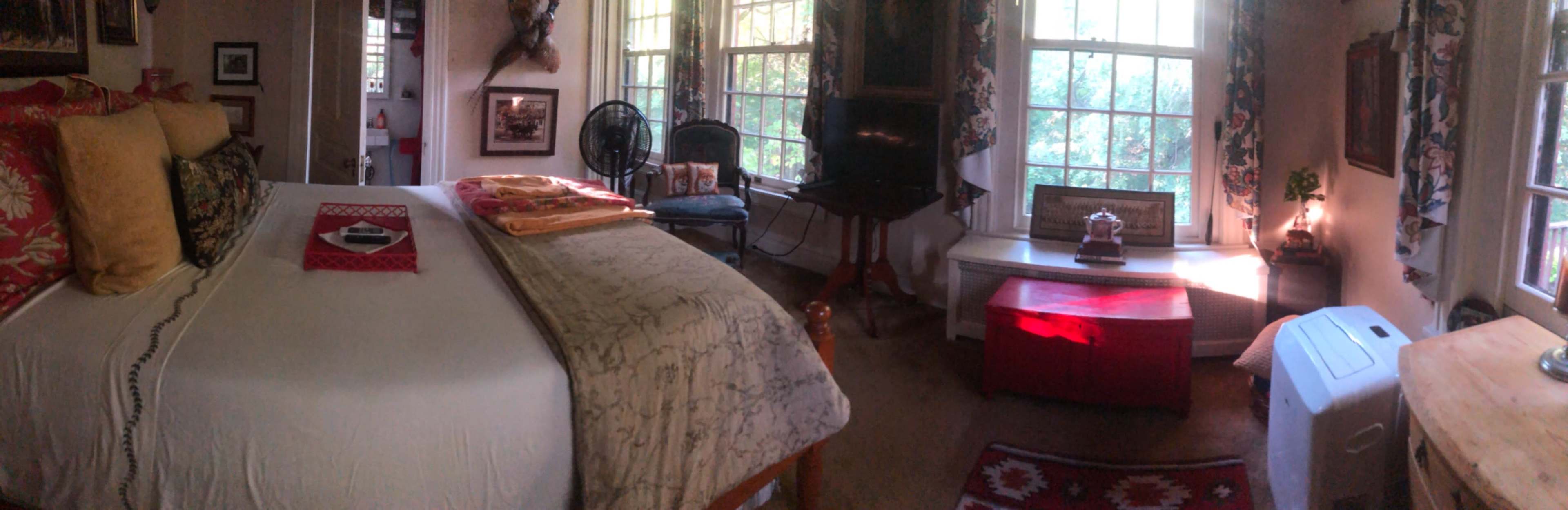 A cozy bedroom with a bed, a fan, a desk, a small red table, and windows allowing natural light to enter.