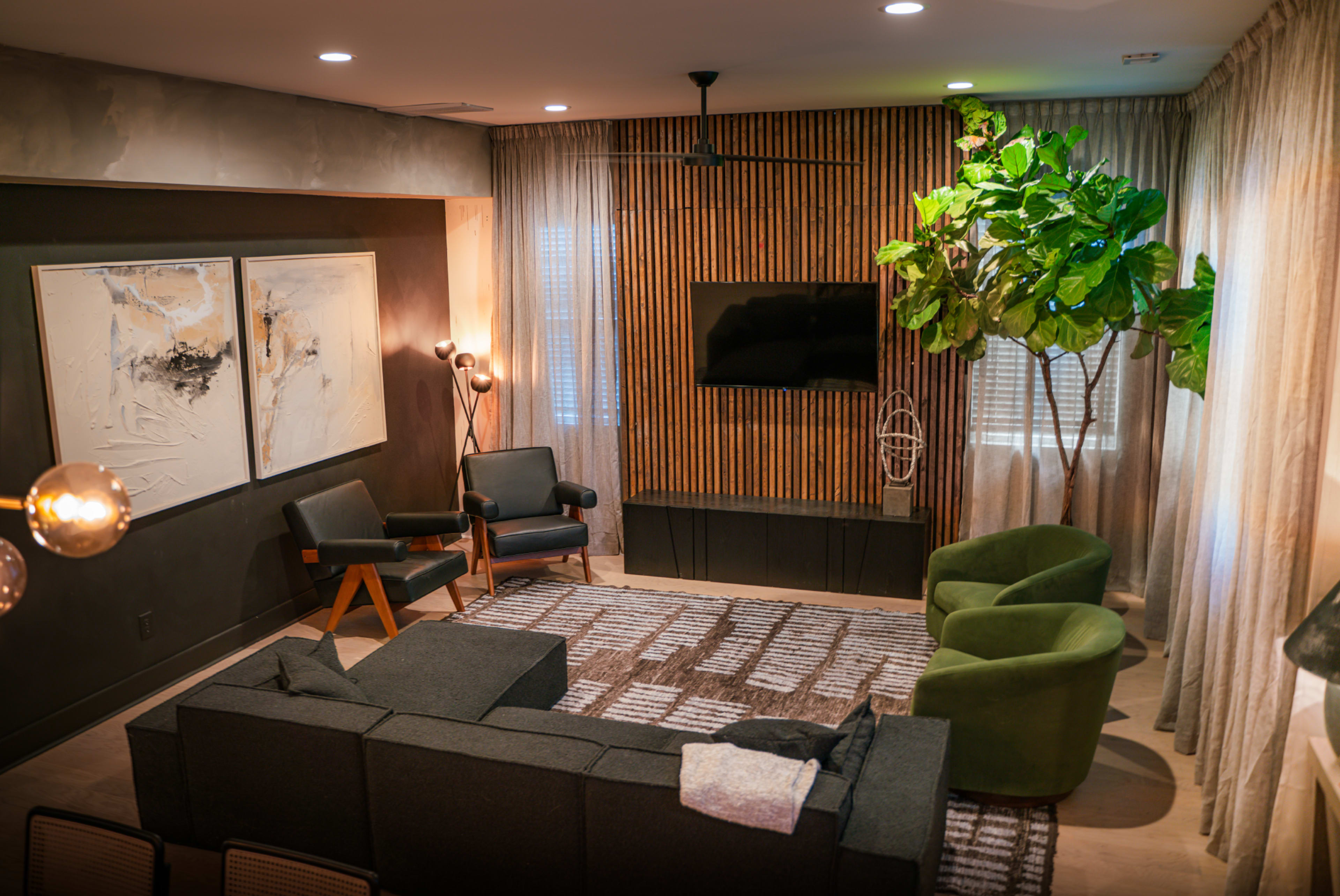 A modern living room featuring a gray sectional sofa, two black armchairs, a green lounge chair, a TV mounted on a wooden panel, and large curtains.