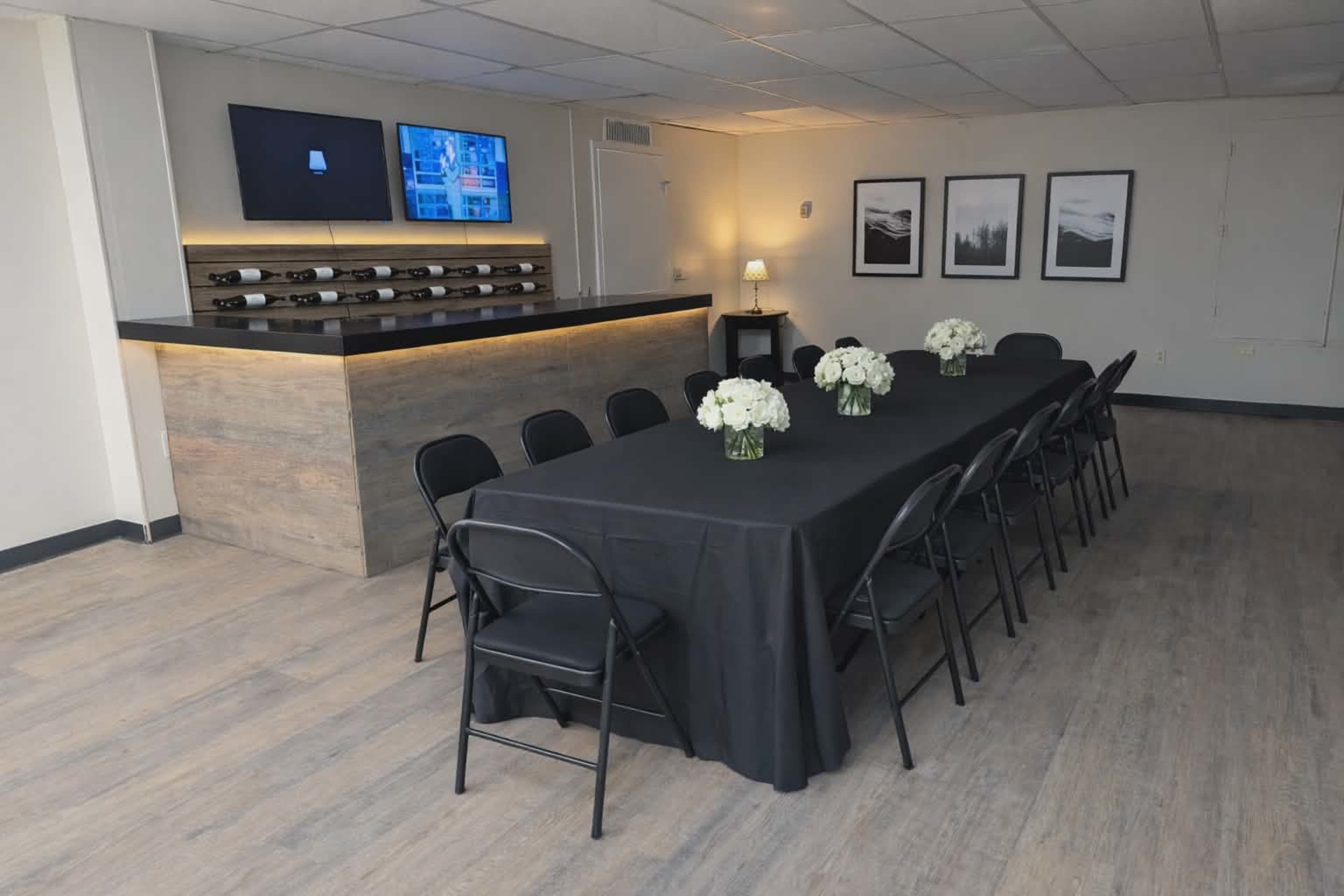 A conference room features a long black table set with floral centerpieces, surrounded by folding chairs, with a bar area and wine storage in the background.