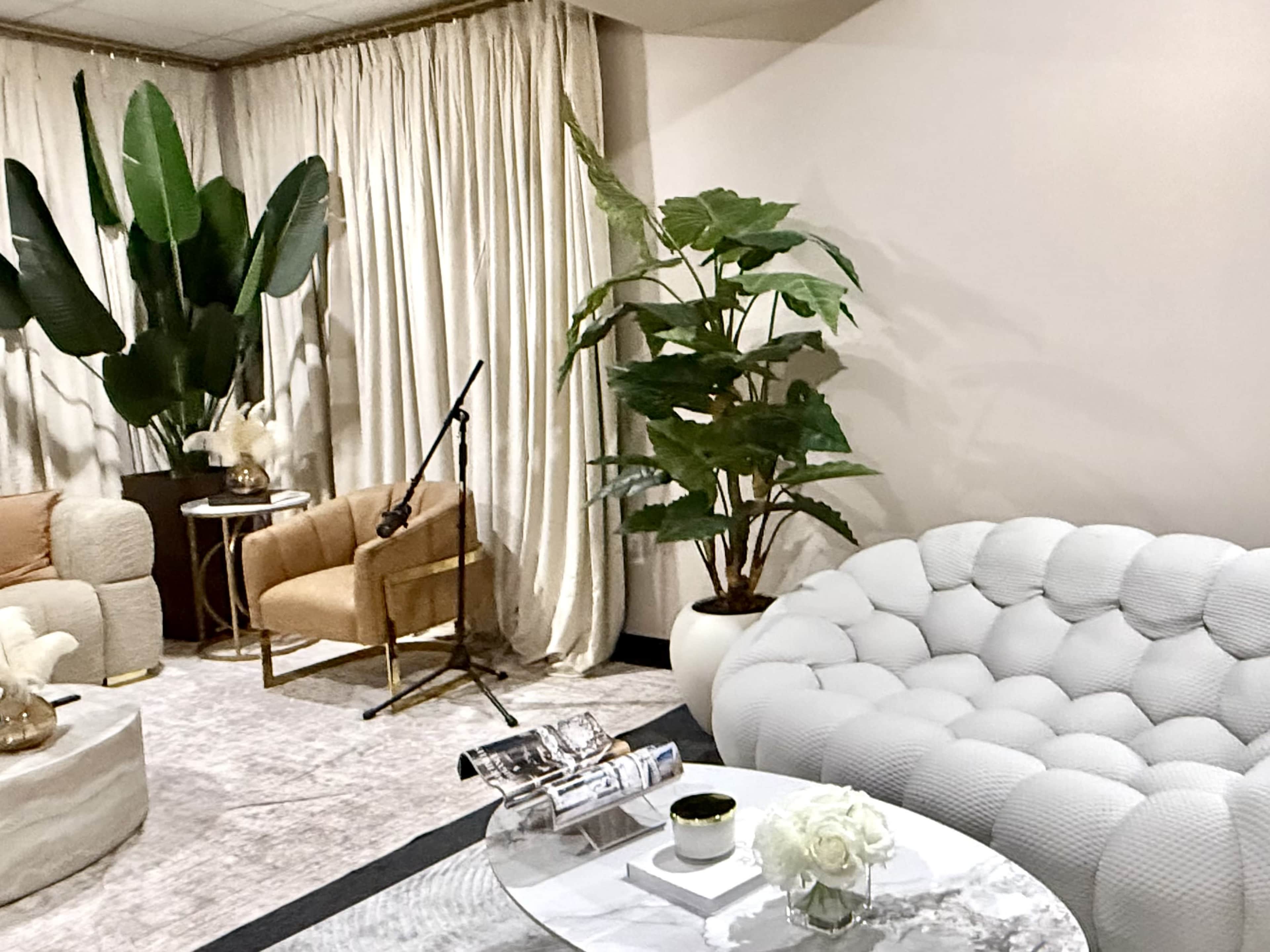 The image shows a modern living space with a white tufted sofa, a round coffee table, and decorative plants amid soft curtains.