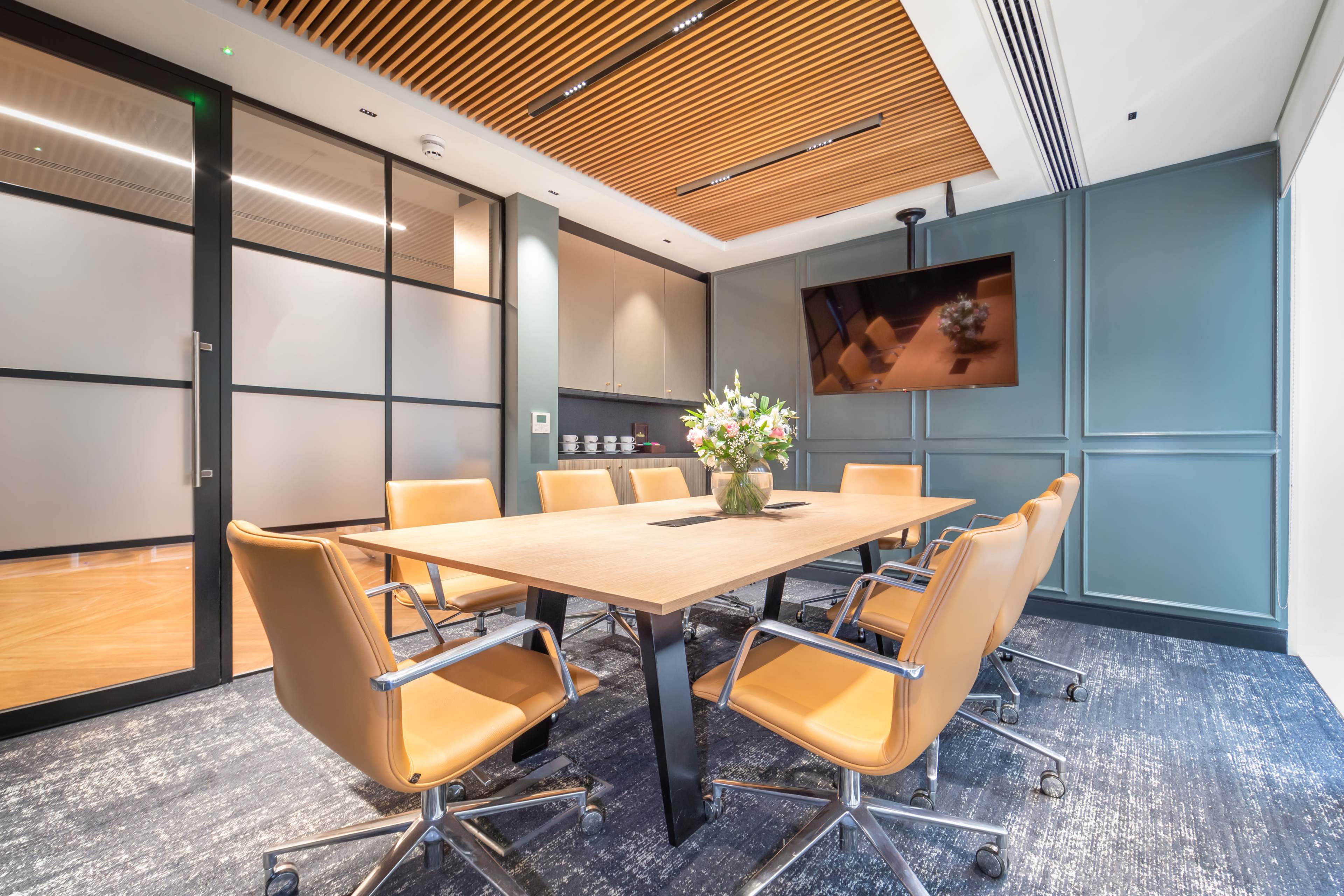 A modern conference room features a large wooden table surrounded by leather chairs, with a wall-mounted television and a floral centerpiece.