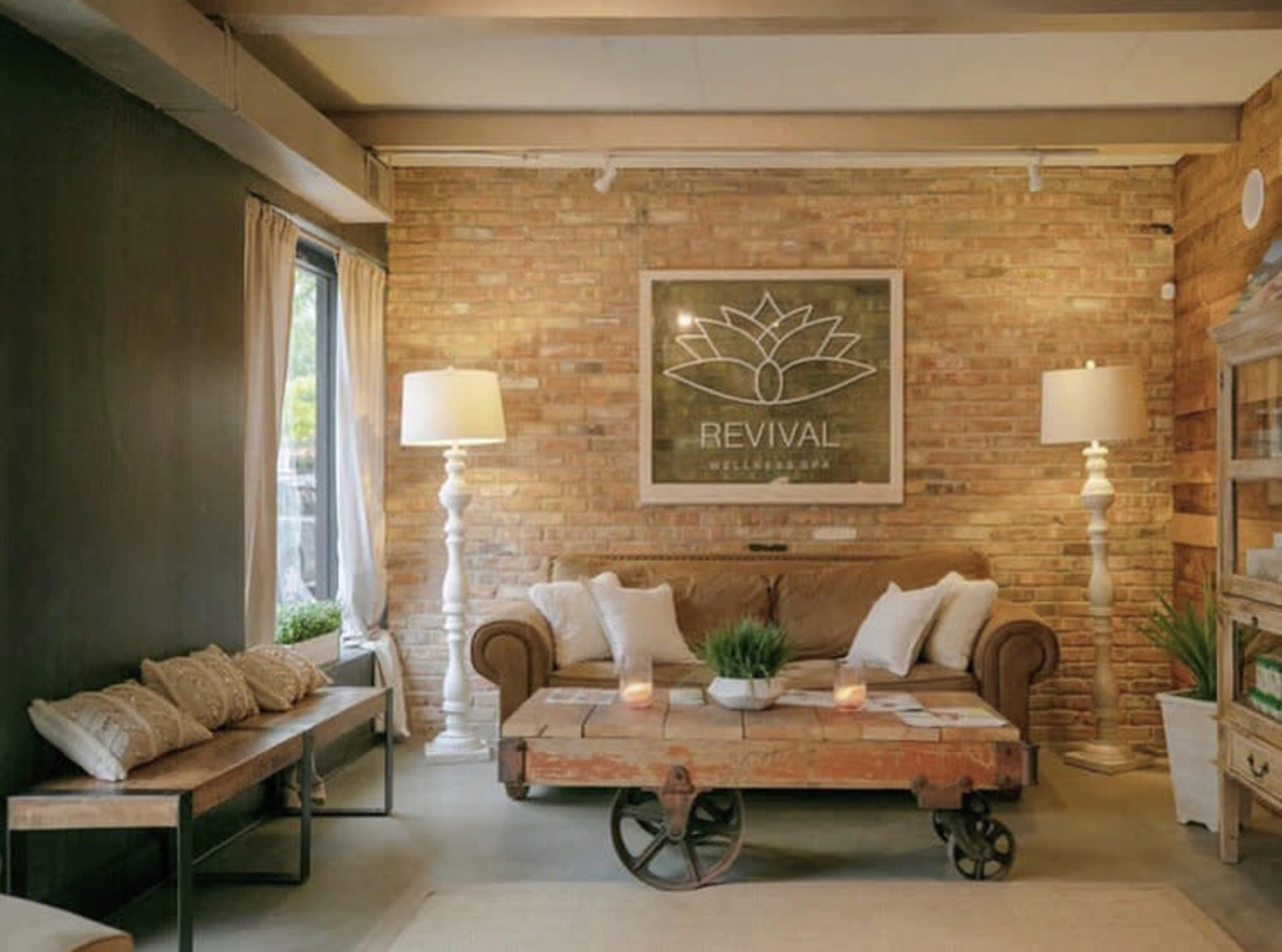 A cozy lounge area featuring a brown sofa, a rustic coffee table on wheels, and decorative lamps against a brick wall with a "Revival Wellness" logo.