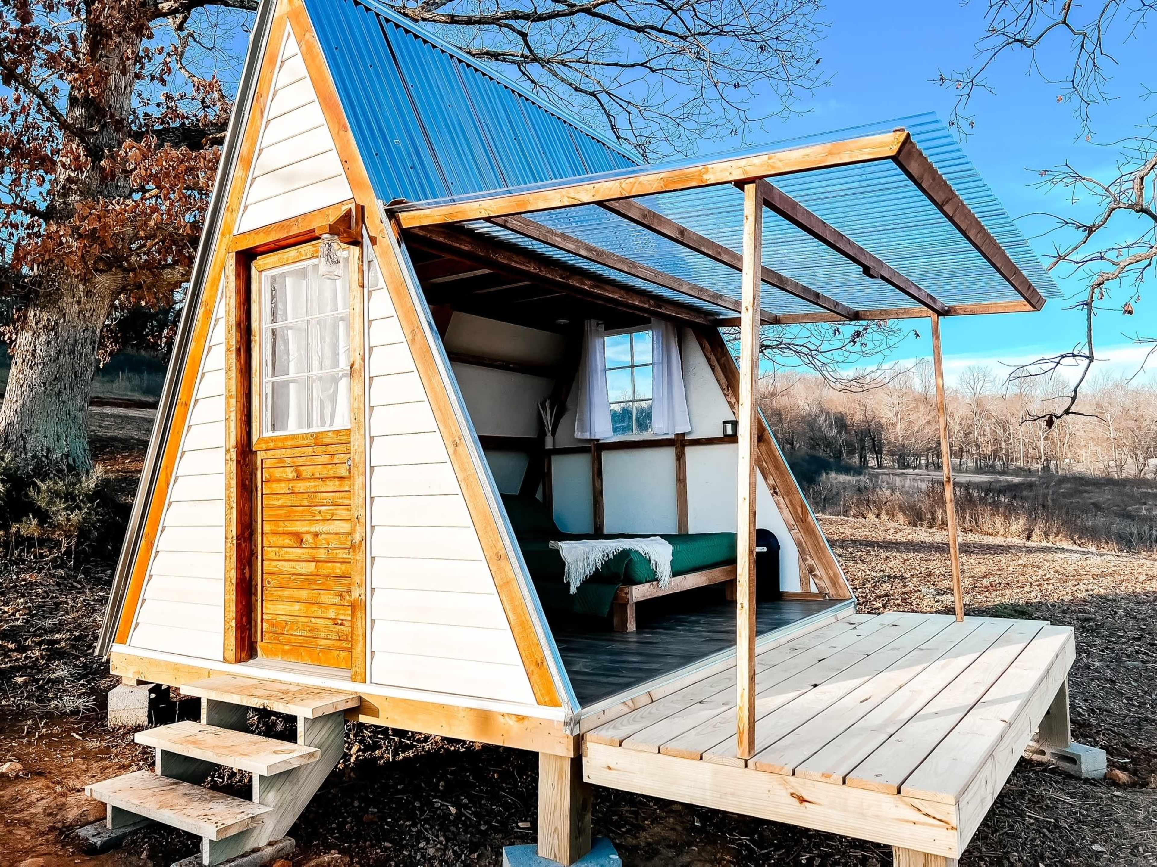 A triangular roof A-frame cabin with a wooden deck and steps, set in a rural landscape.
