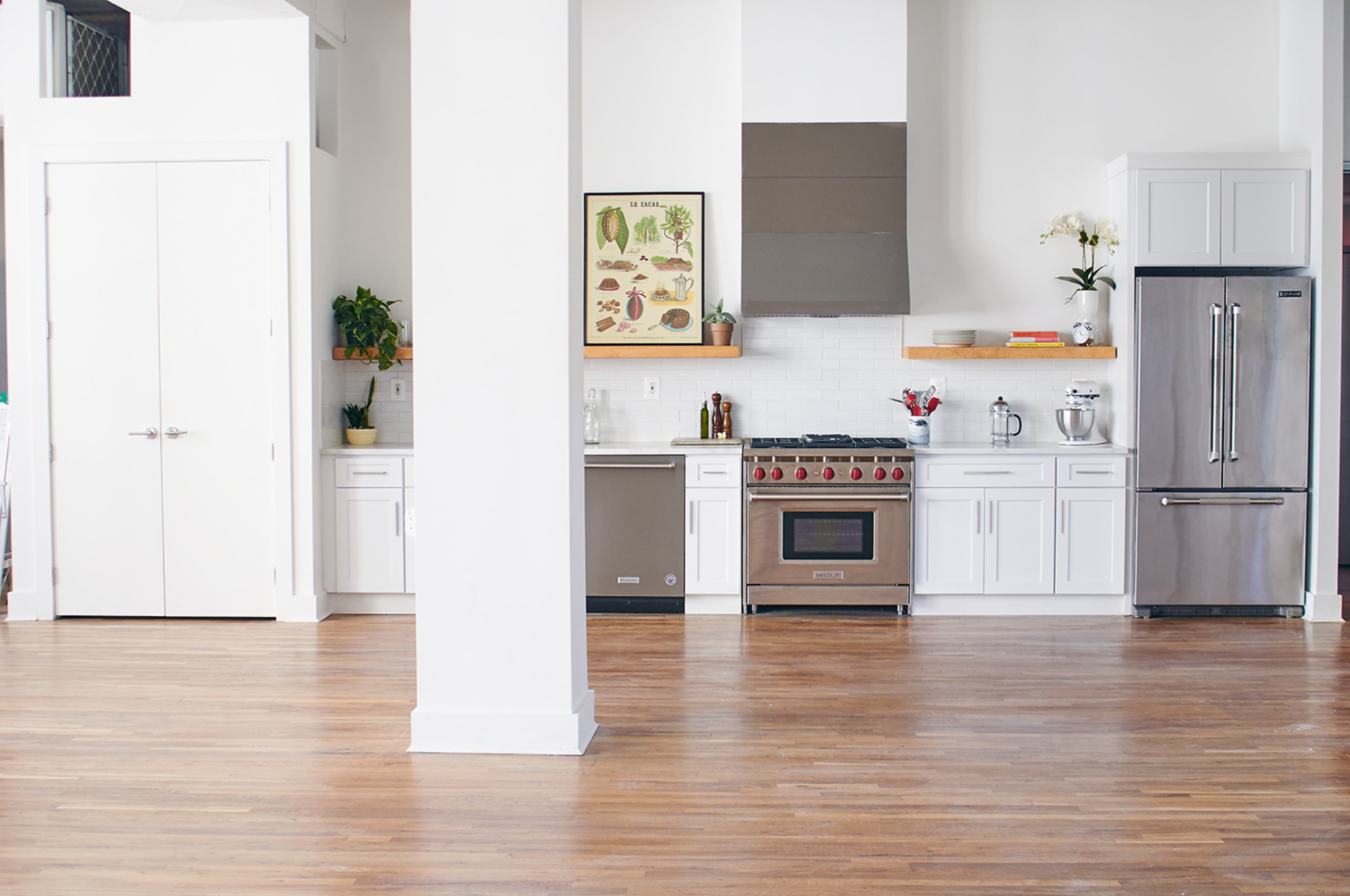 A modern kitchen featuring white cabinets, a stainless steel range and refrigerator, and wooden shelves with decorative plants, all set in a spacious room with hardwood flooring.