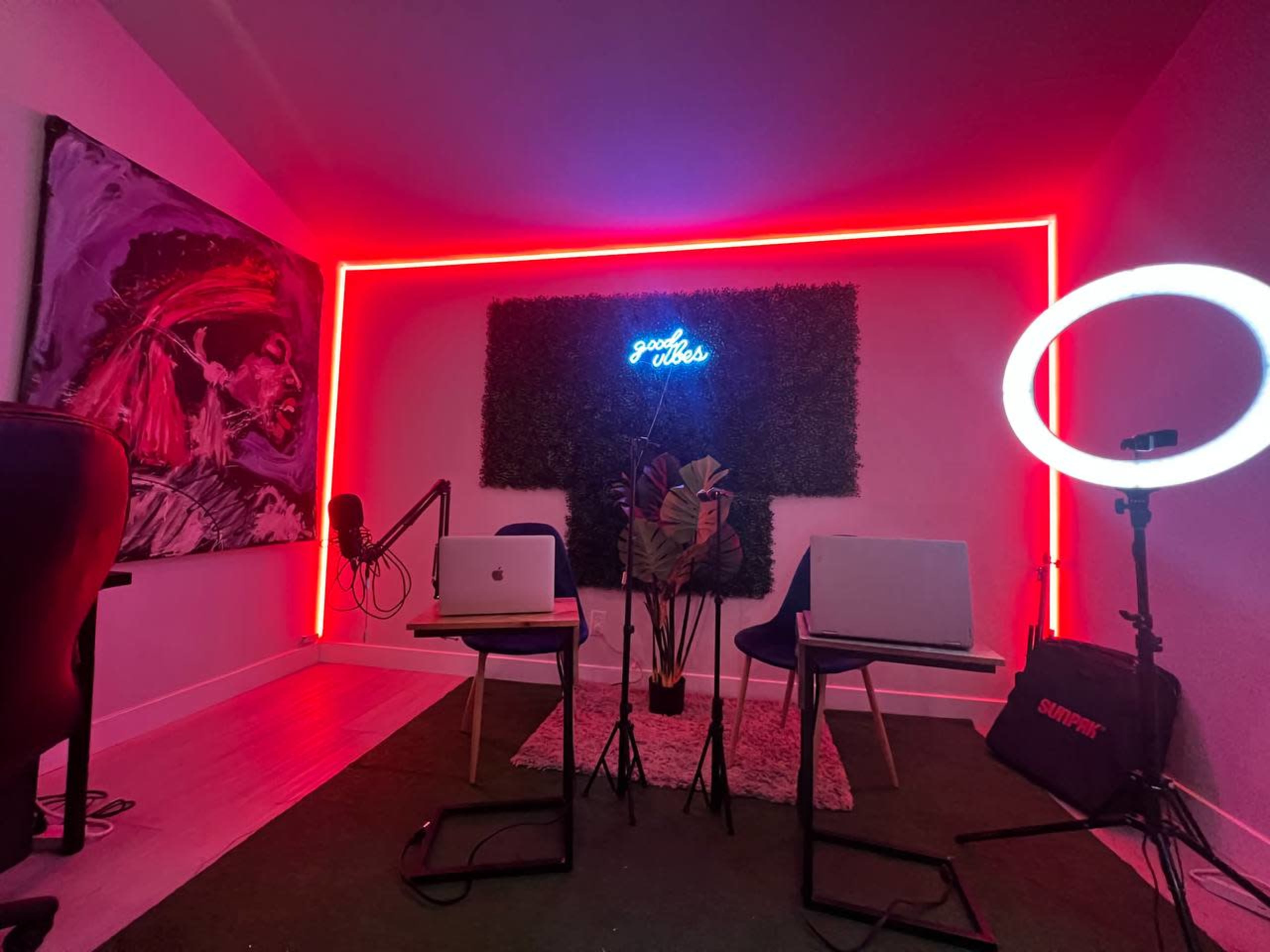 A stylish room setup for a podcast, featuring neon lighting, two laptops on tables, and a ring light next to a microphone.