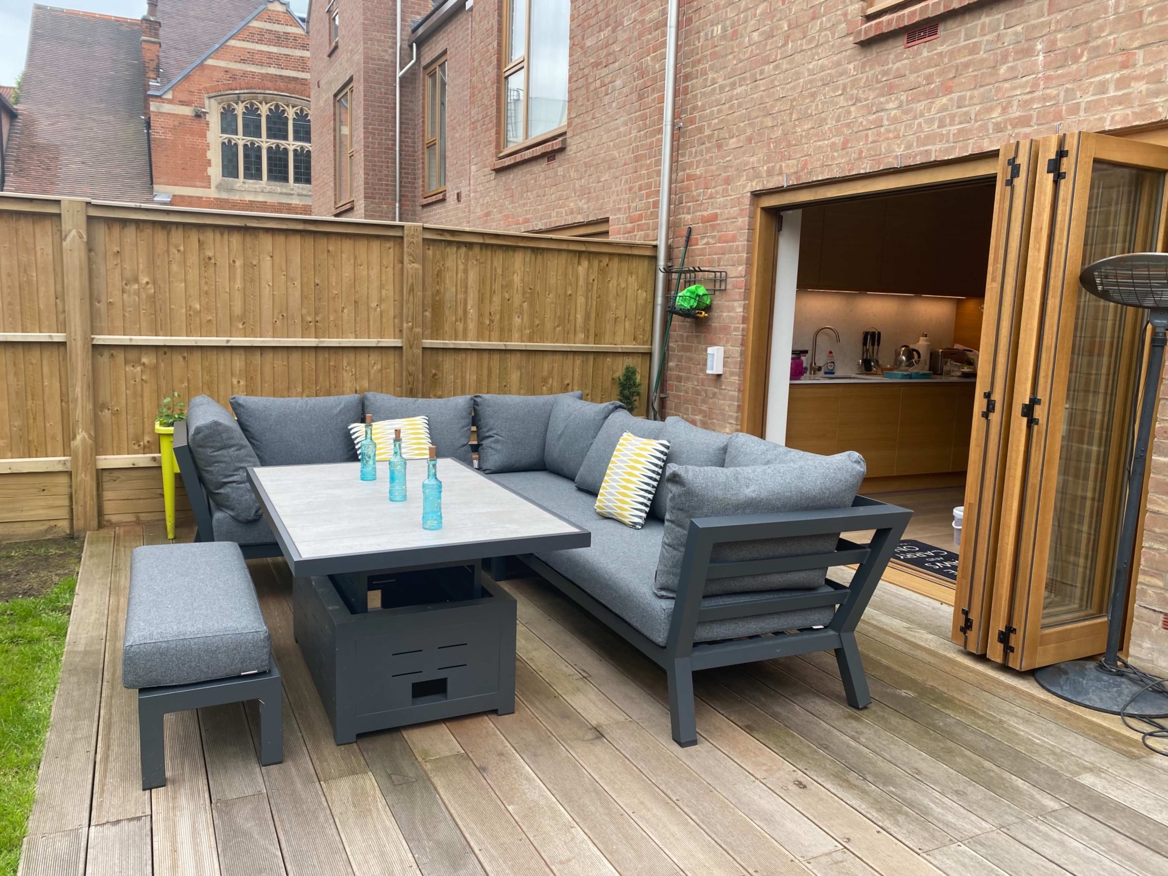 A patio area features a large gray sectional sofa with a matching table and several decorative cushions, surrounded by wooden fencing and leading to an open door into a kitchen.