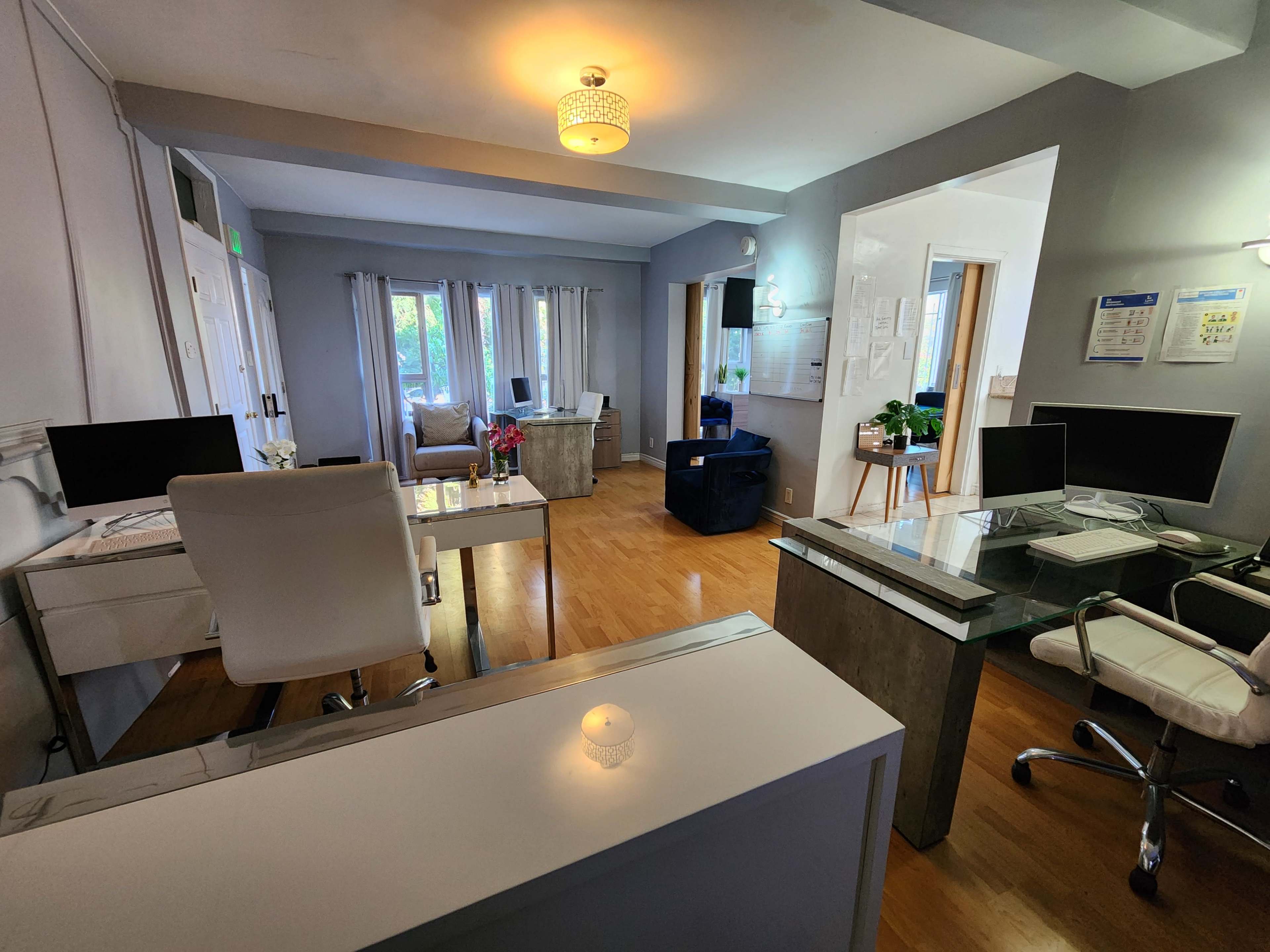 The image shows an office space with two desks, computer setups, a seating area, and natural light coming through the windows.