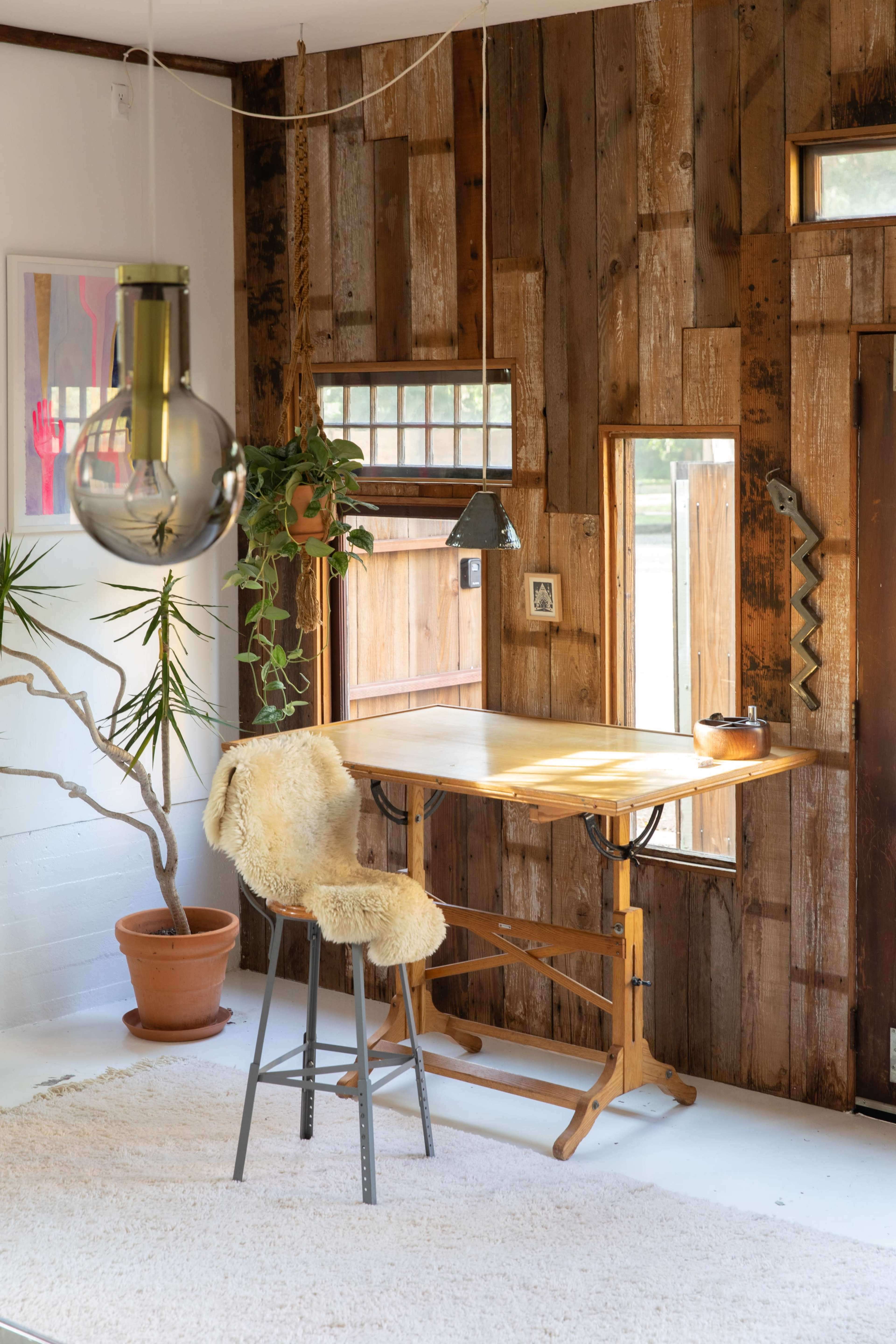 A wooden table with a high stool is positioned in a room featuring rustic wooden walls, a plant in a pot, and a hanging light fixture.