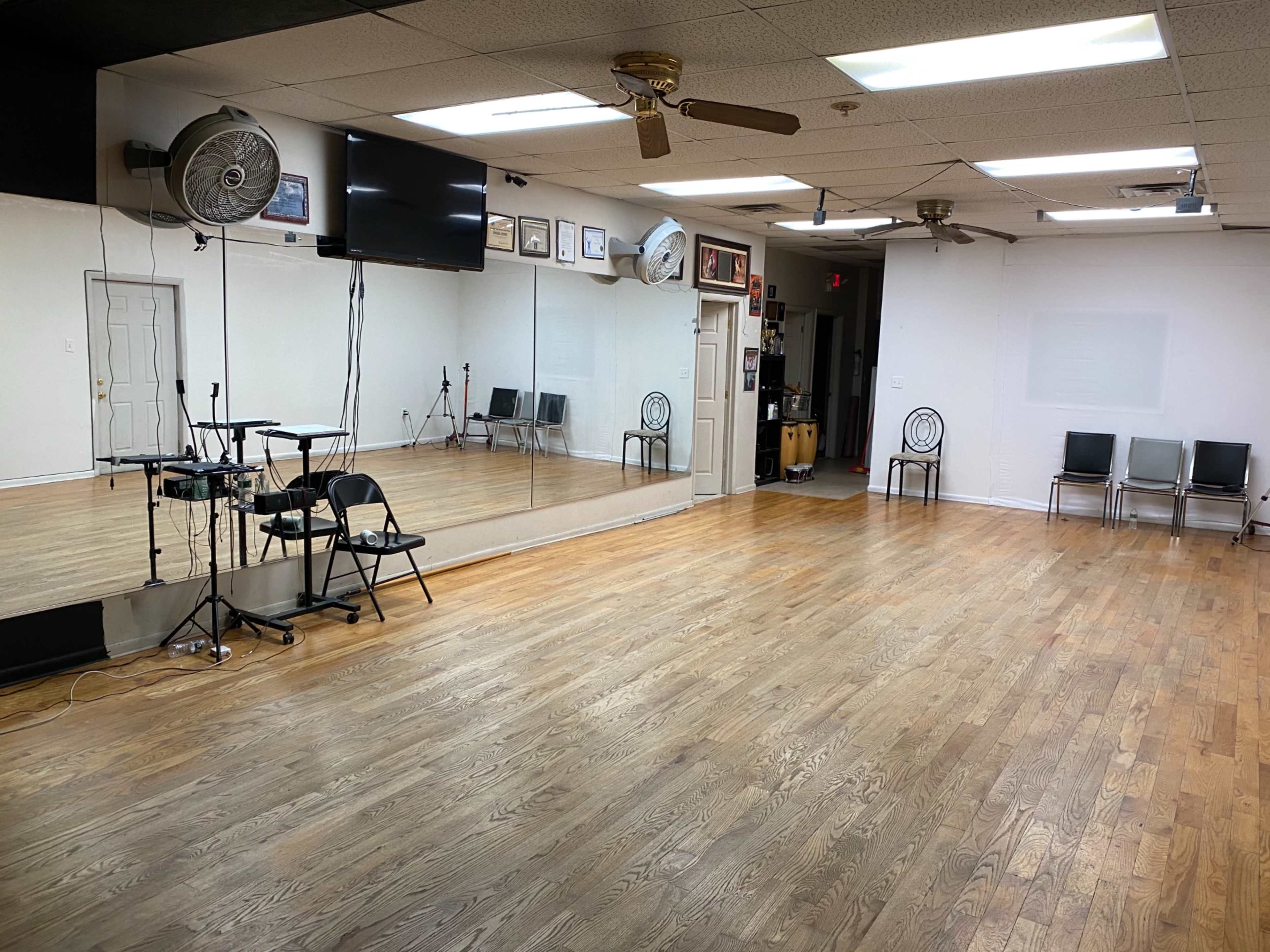 An empty dance studio featuring wooden floors, mirrors on one wall, and multiple chairs arranged on the opposite side.