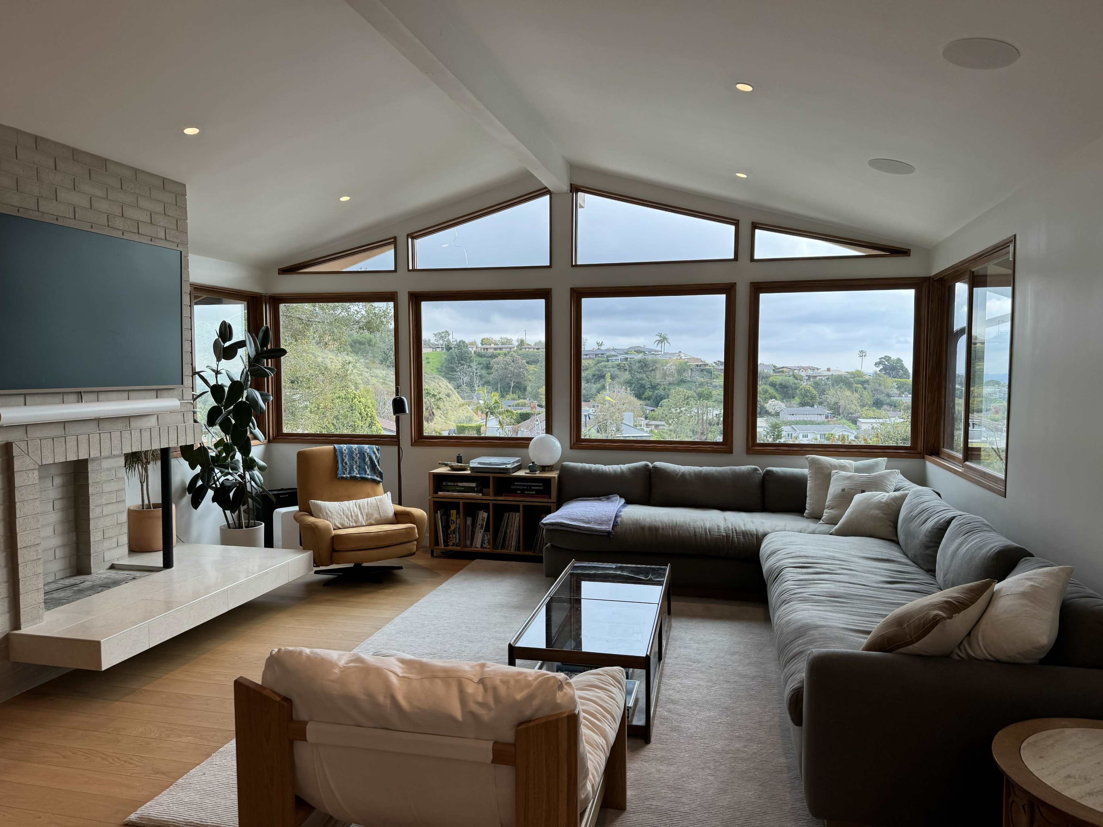 The modern living room features large windows that offer a view of the outdoors, with a sectional sofa, an armchair, a coffee table, and a fireplace.