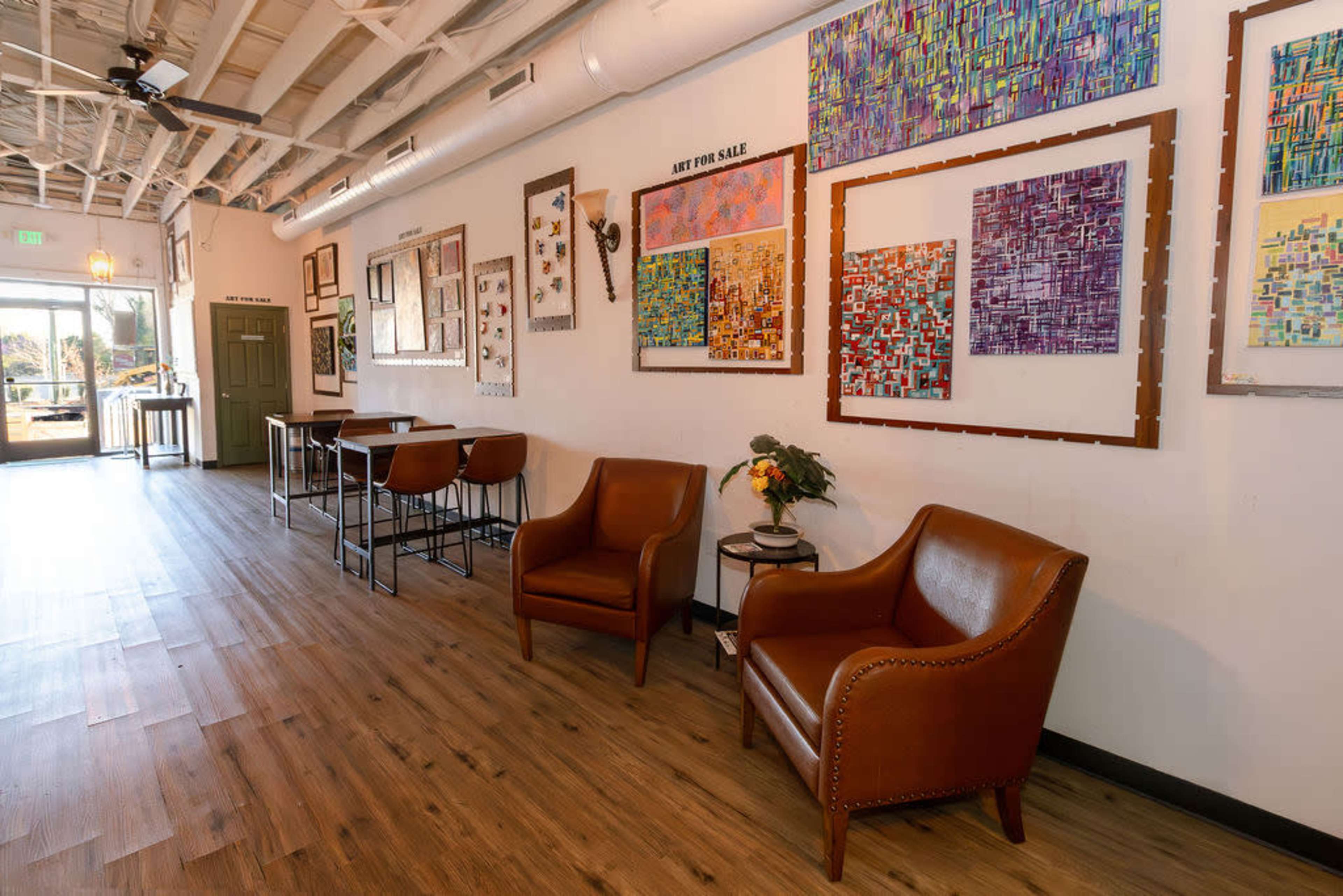 The image shows a well-lit indoor space featuring two brown leather armchairs, a small table with a flower arrangement, and several colorful artworks displayed on the wall.