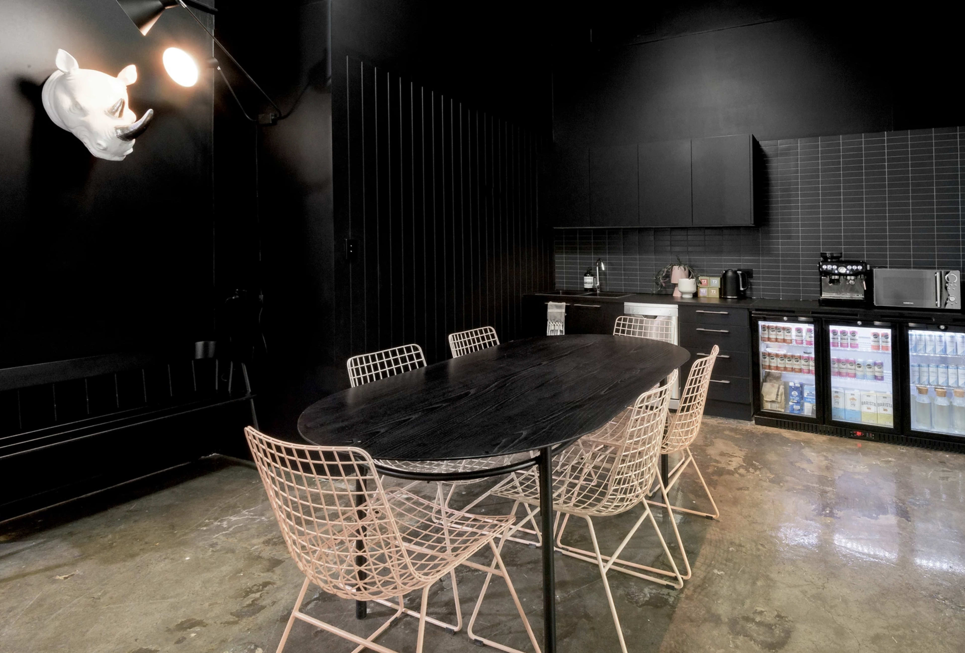 The image shows a modern kitchen space with a large black table surrounded by wire chairs, featuring dark walls and a sleek counter with appliances and a mini fridge.
