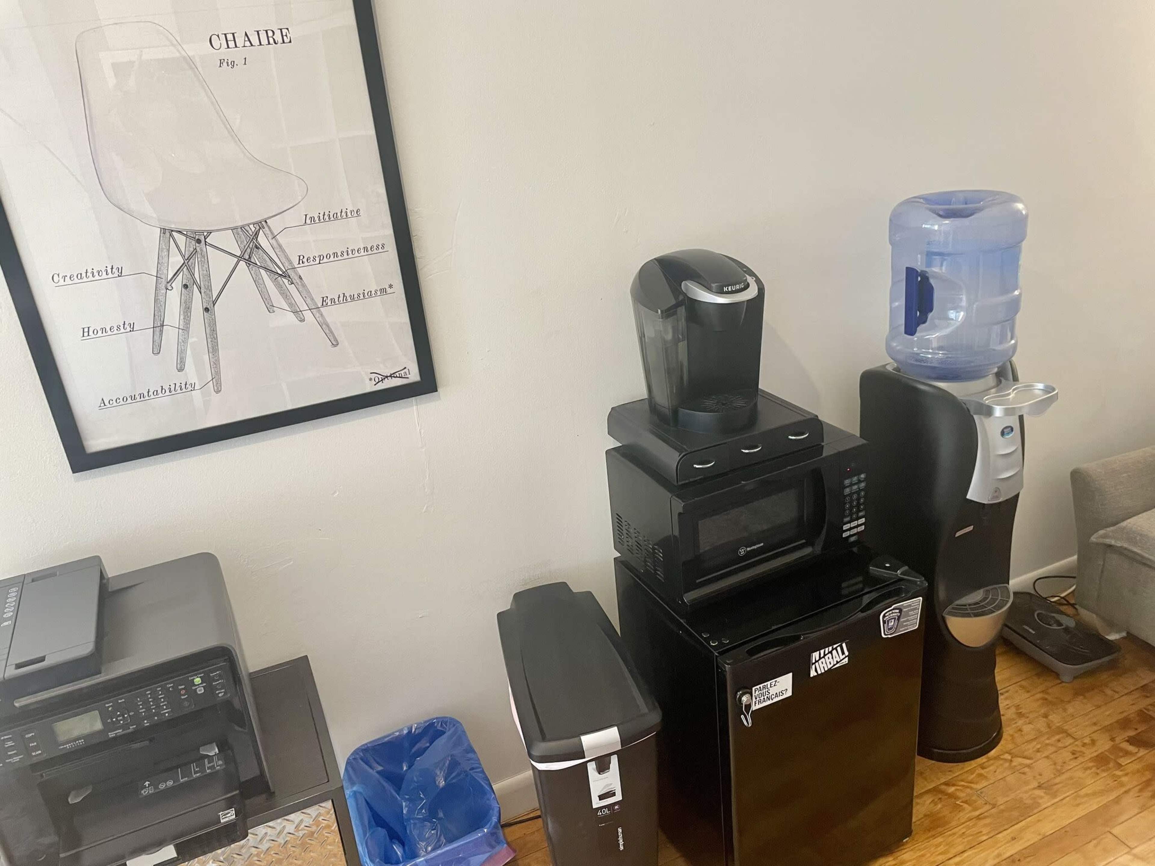 The image shows a corner of a room featuring a wall poster, a coffee maker, a microwave, a small refrigerator, a trash bin, a recycling bin, and a water cooler.