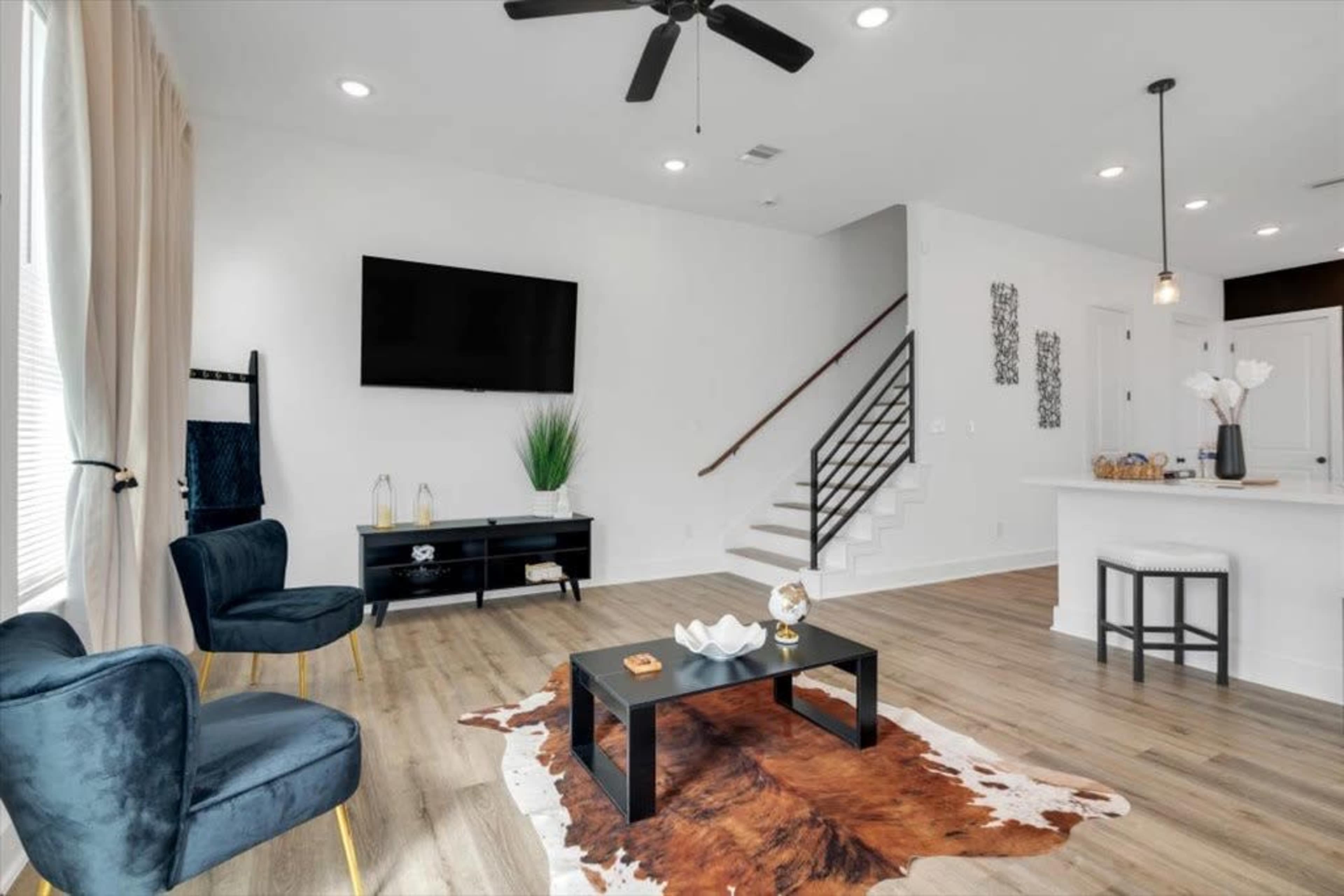 The living room features a mounted TV, a coffee table on a cowhide rug, two blue chairs, and a staircase leading to an upper level.